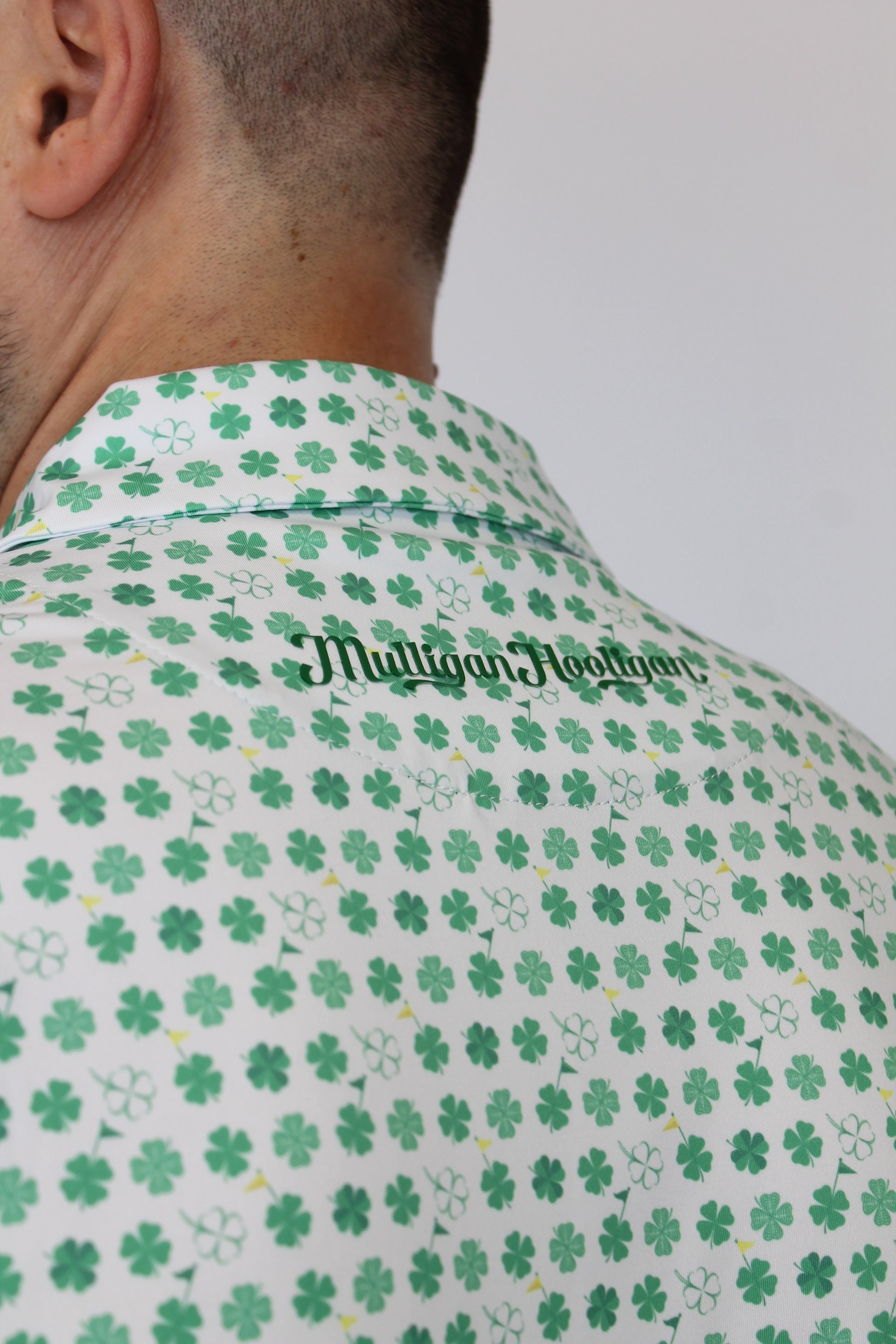 Close-up of a person wearing a white golf polo with green four-leaf clover pattern and 'Mulligan Hooligan' printed vertically in green script.