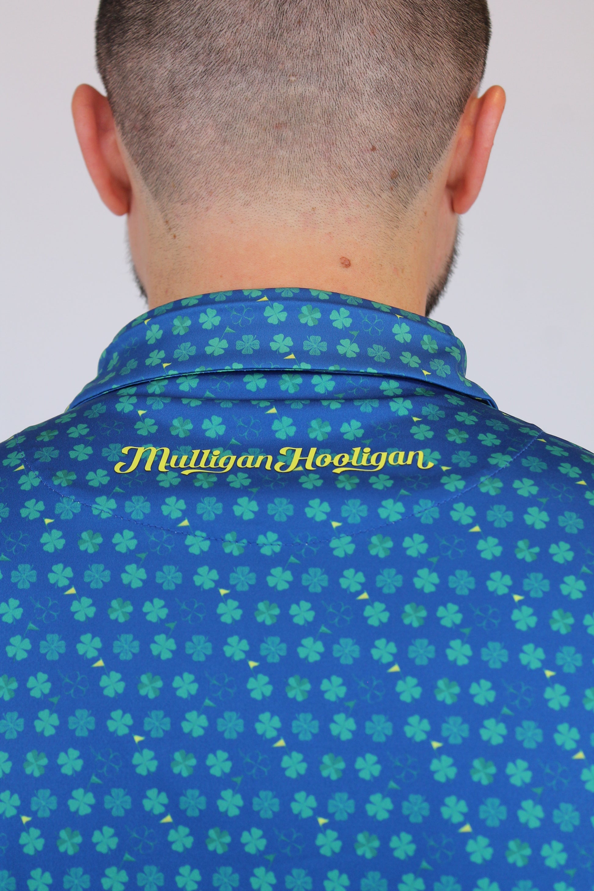 Close-up back view of a person wearing a blue 4-leaves Clover patterned collared shirt with green clover motifs and yellow 'Mulligan Hooligan' script on the collar.