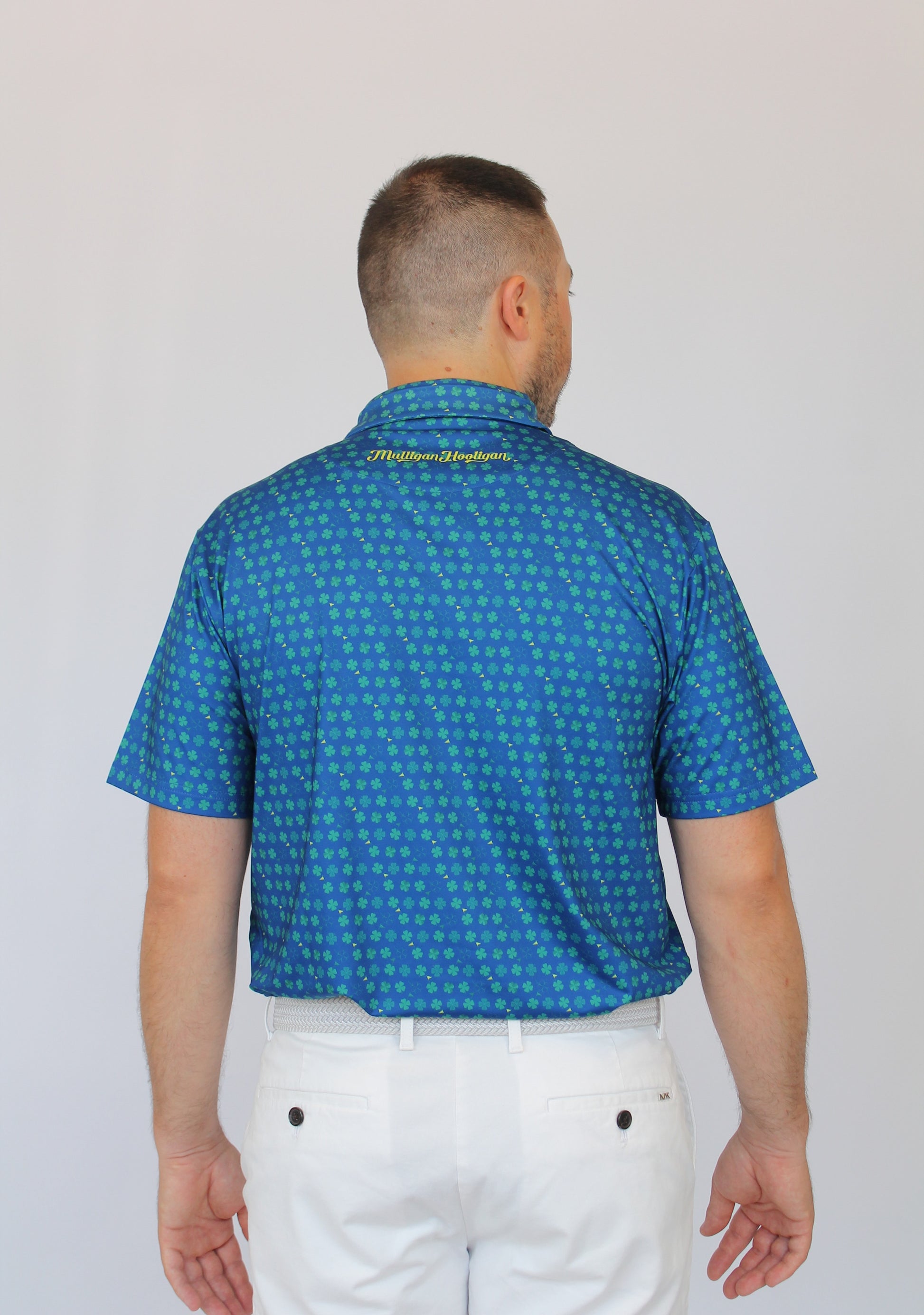 A man viewed from behind wearing a blue short-sleeve Clover patterned golf shirt and white pants against a plain light gray background.