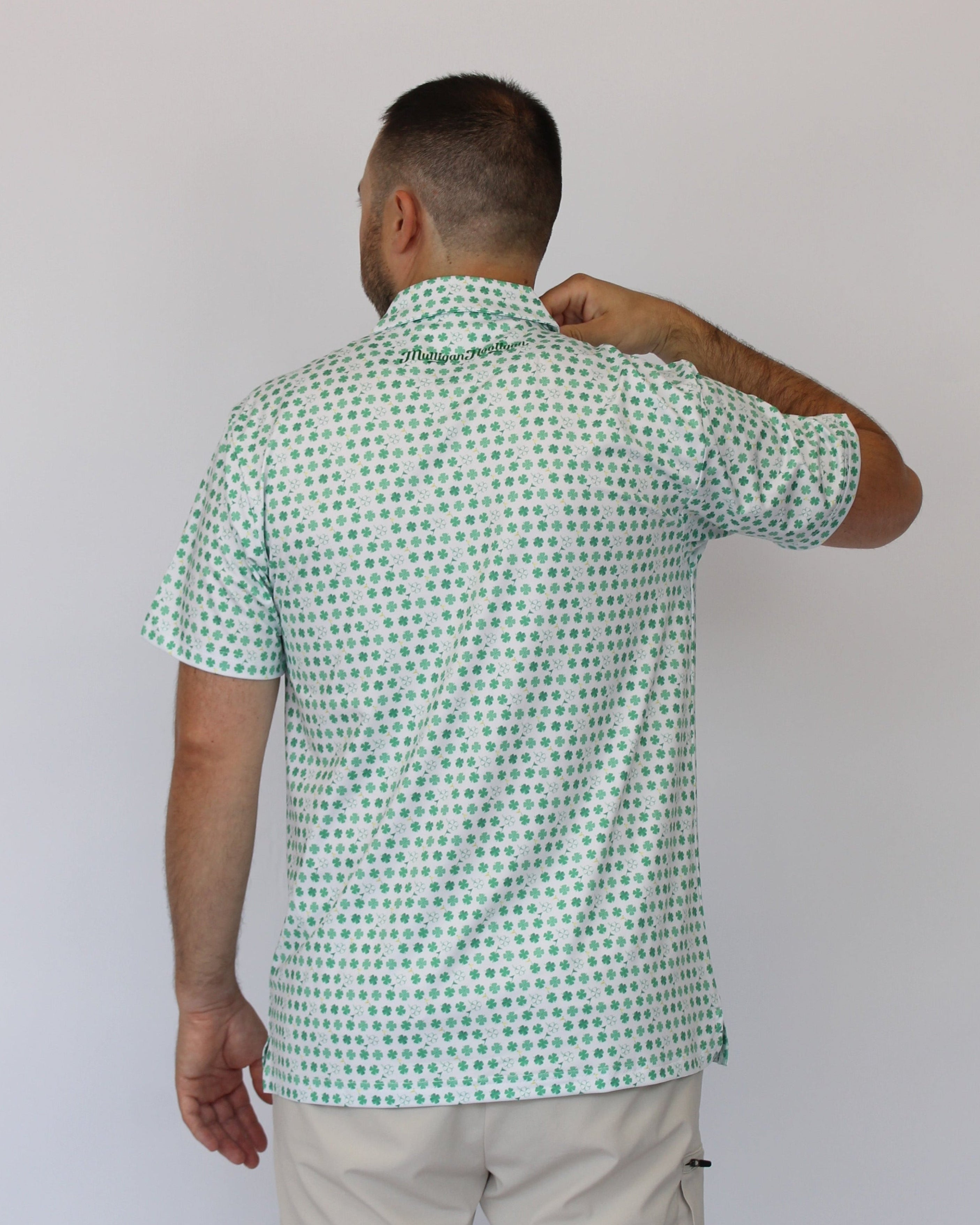 A man viewed from behind wearing a short-sleeve button-up shirt with a green and white clover pattern, paired with light beige pants, against a plain light gray background.