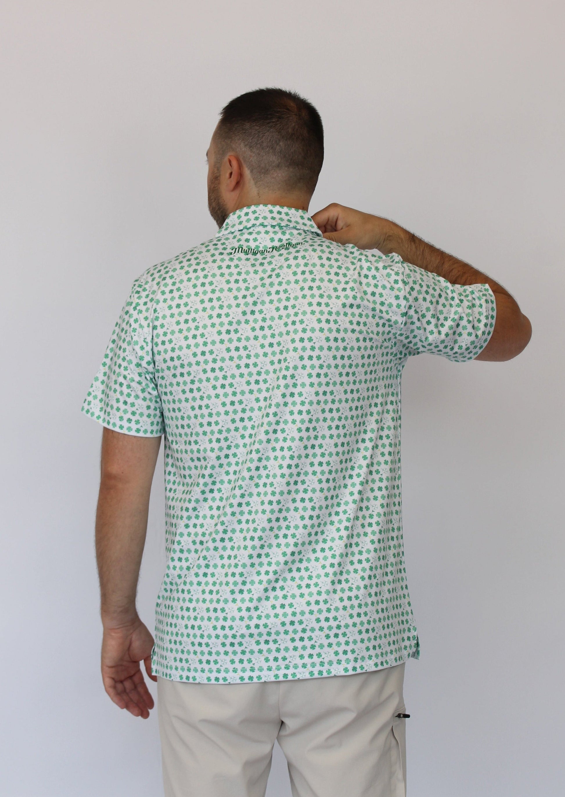 A man viewed from behind wearing a short-sleeve button-up shirt with a green and white clover pattern, paired with light beige pants, against a plain light gray background.