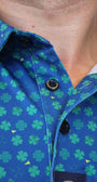 Close-up of a blue button-up shirt with a green shamrock pattern, worn by a person. The shirt features black snap buttons and a visible brand label on the sleeve.