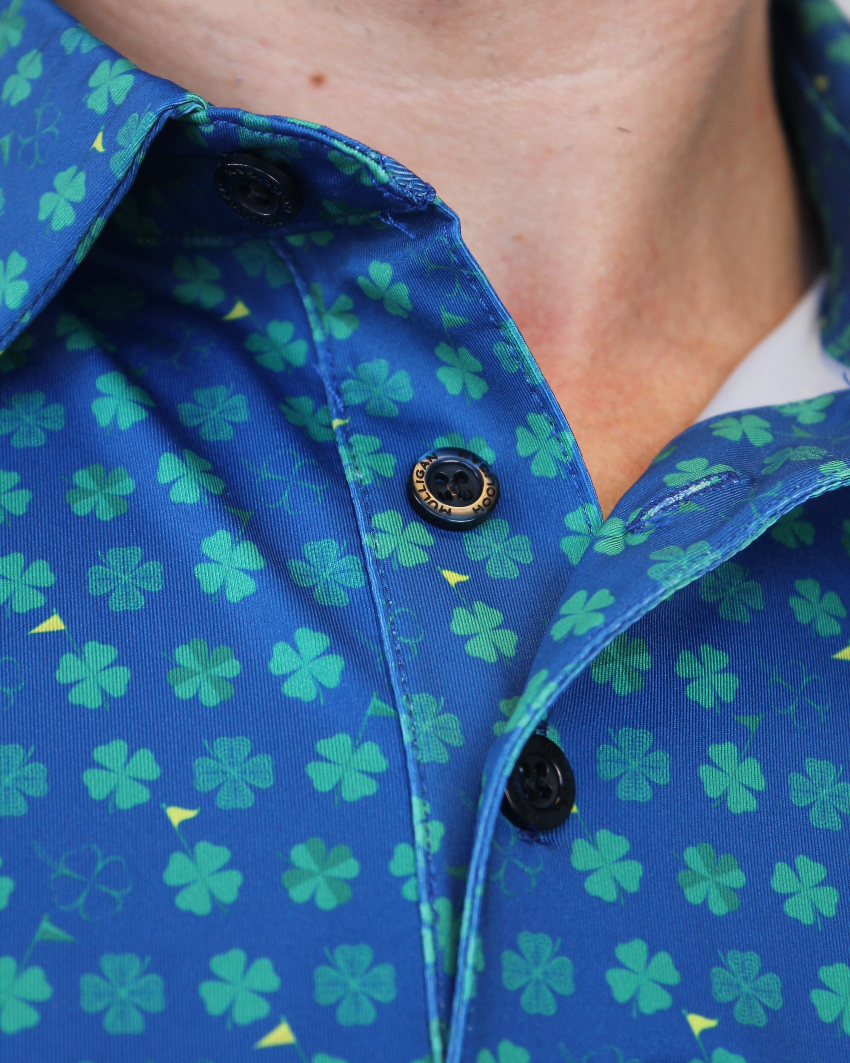 Close-up of a blue button-up shirt with a green shamrock pattern, worn by a person. The shirt features black snap buttons and a visible brand label on the sleeve.