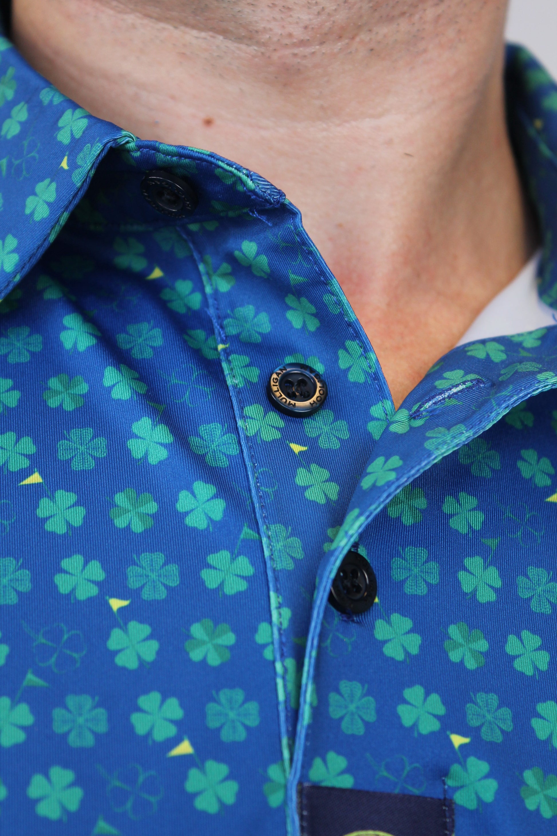 Close-up of a blue button-up shirt with a green shamrock pattern, worn by a person. The shirt features black snap buttons and a visible brand label on the sleeve.