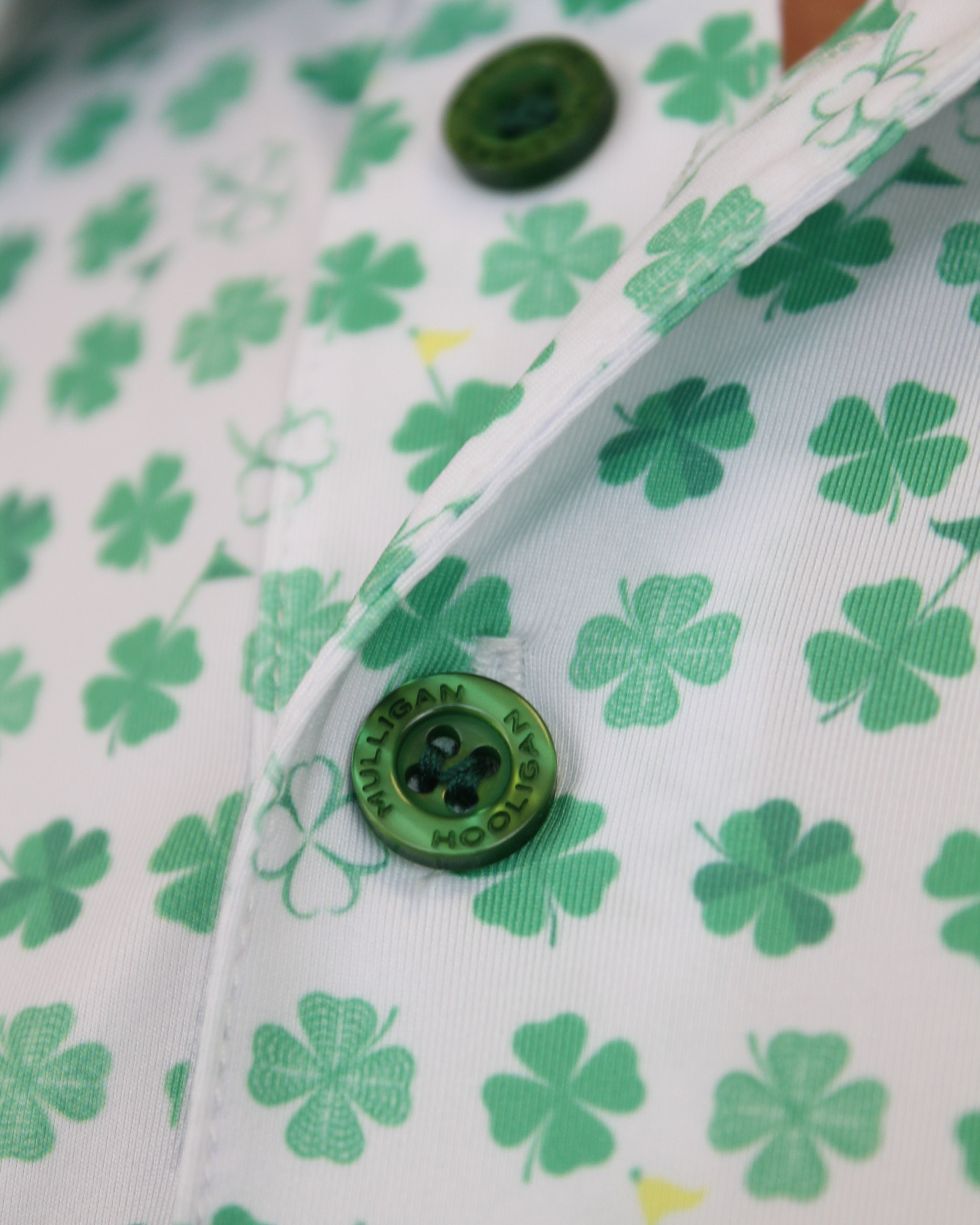 Close-up of a white button-up shirt with a green shamrock and clover pattern, featuring a green button with 'Mulligan Hooligan' text.