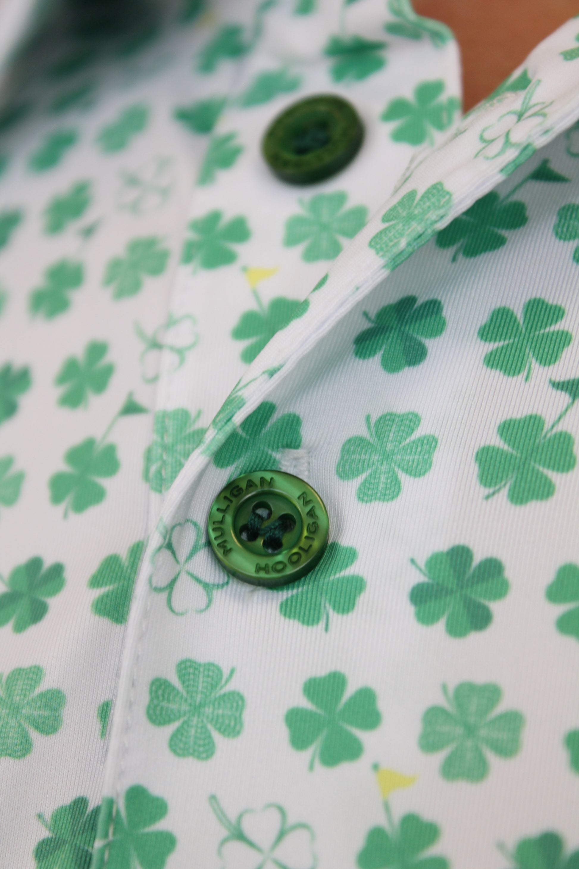 Close-up of a white button-up shirt with a green shamrock and clover pattern, featuring a green button with 'Mulligan Hooligan' text.