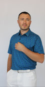 A man wearing a blue short-sleeve button-up shirt with a small repeating clover pattern, paired with white trousers and a white belt, standing against a plain light gray background.