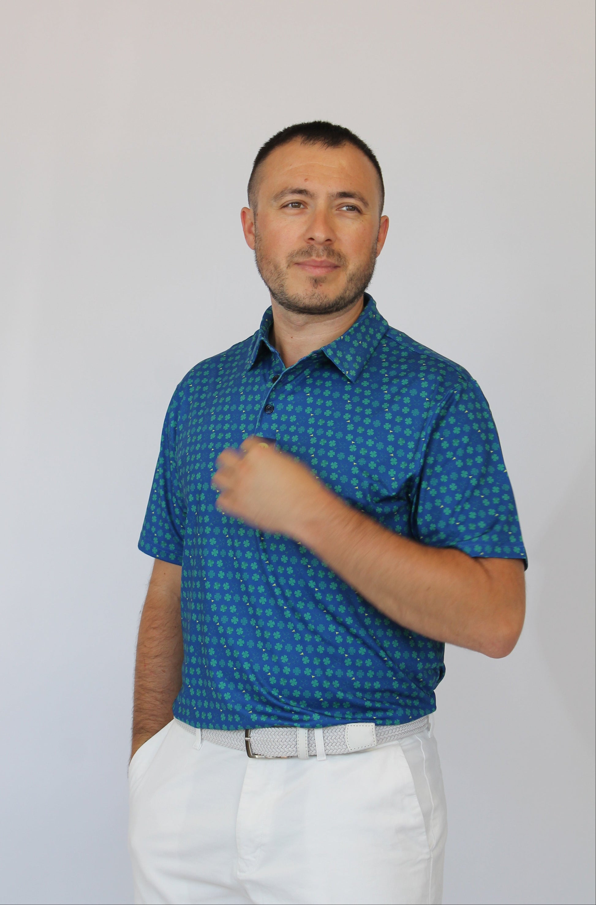 A man wearing a blue short-sleeve button-up shirt with a small repeating clover pattern, paired with white trousers and a white belt, standing against a plain light gray background.