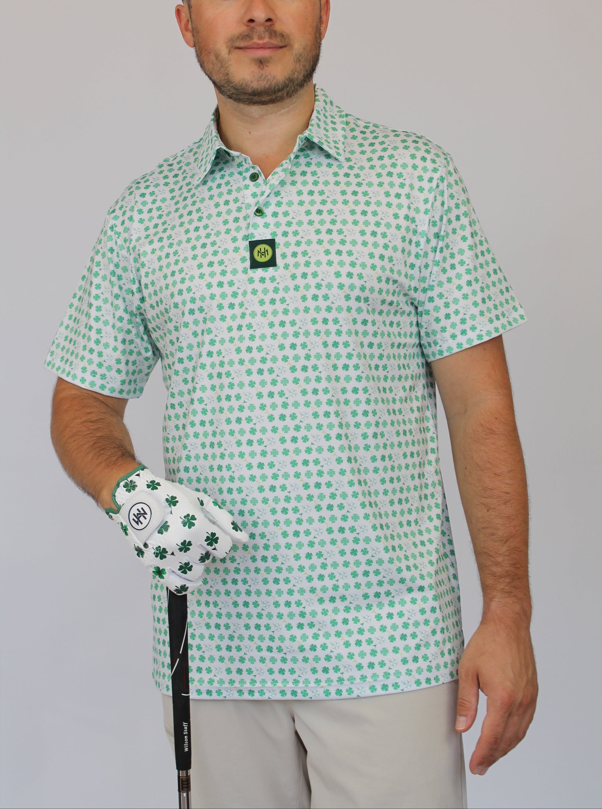A man wearing a short-sleeve polo shirt with a green and white shamrock pattern, holding a golf club and wearing a matching white golf glove with green shamrocks. The background is a solid light gray.