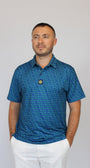 A man wearing a blue short-sleeve polo shirt with a small Clover pattern and white pants, standing against a plain light gray background.
