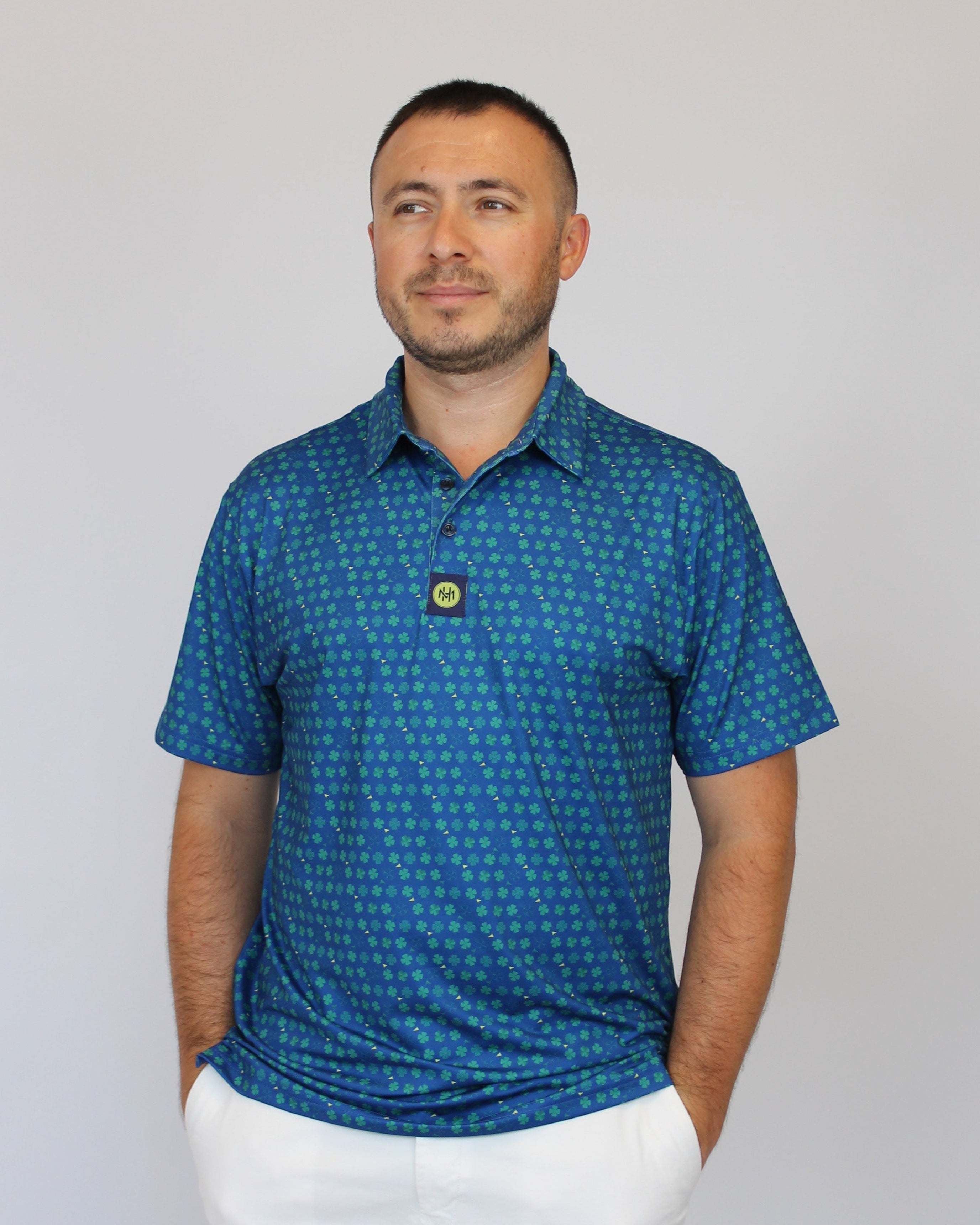 A man wearing a blue short-sleeve polo shirt with a small Clover pattern and white pants, standing against a plain light gray background.