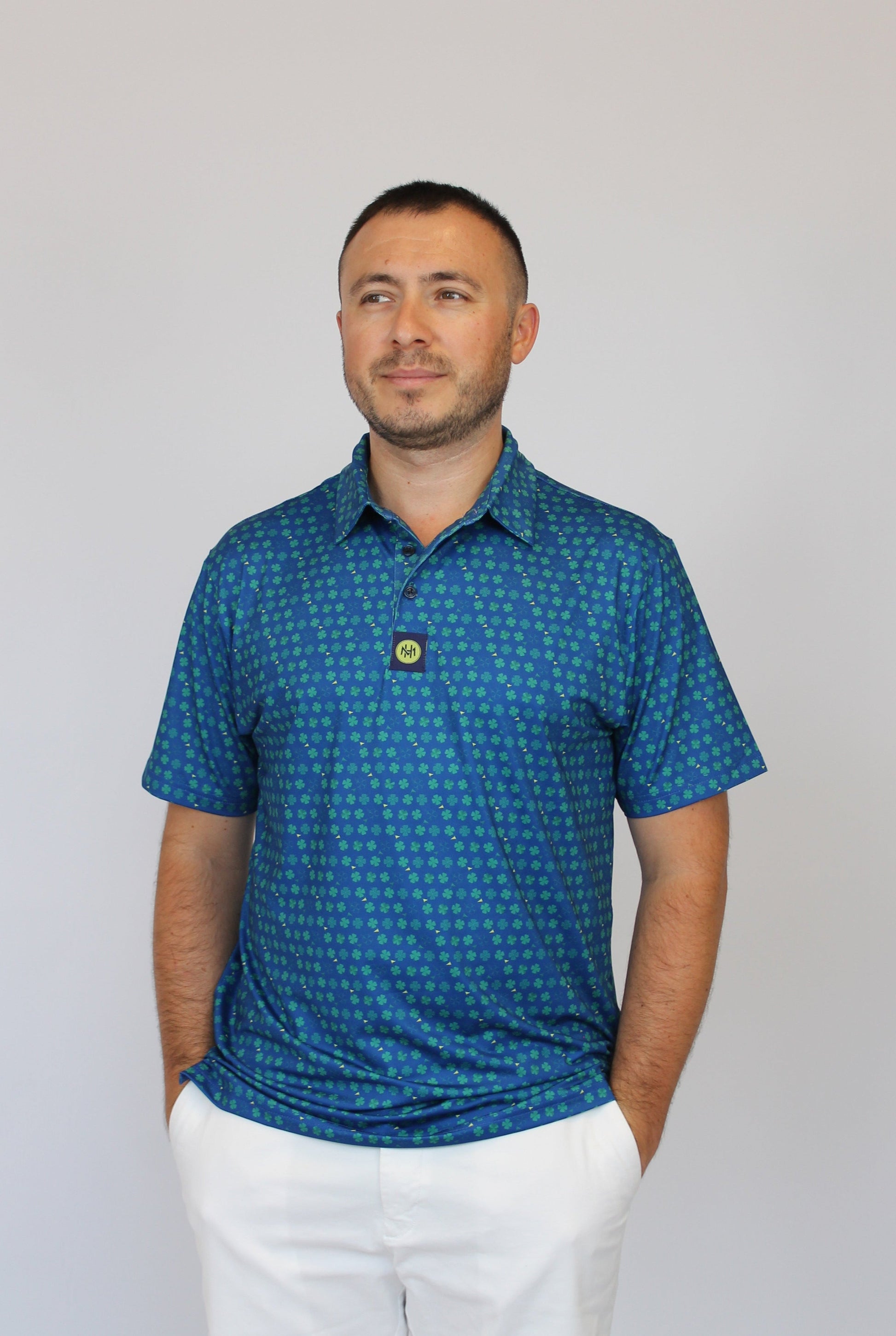 A man wearing a blue short-sleeve polo shirt with a small Clover pattern and white pants, standing against a plain light gray background.
