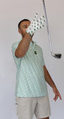 A man wearing a Clover green and white patterned polo shirt and khaki shorts, wearing a Clover Luck golf glove with green Clover, holding a golf club against a plain light gray background.