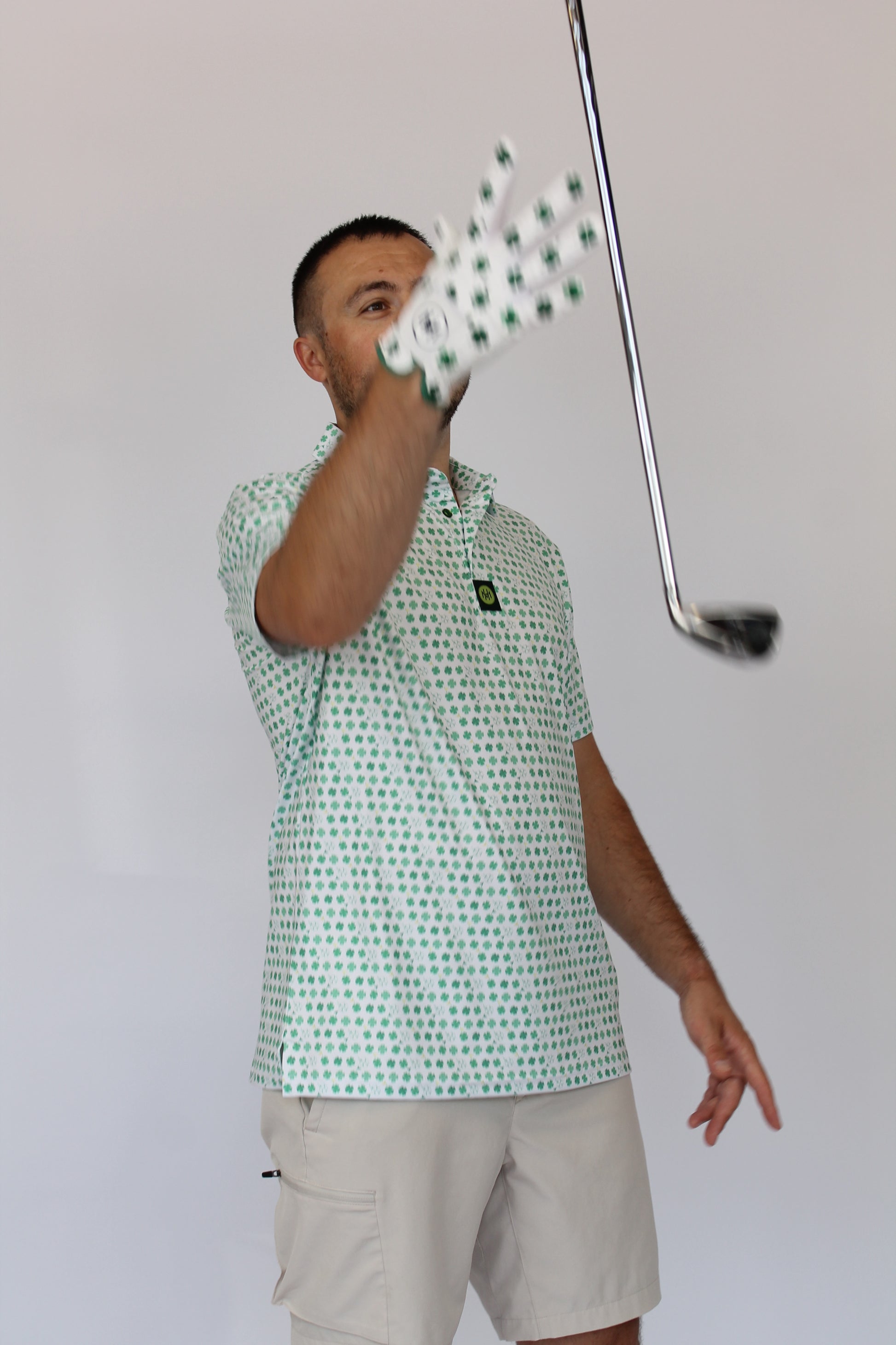 A man wearing a Clover green and white patterned polo shirt and khaki shorts, wearing a Clover Luck golf glove with green Clover, holding a golf club against a plain light gray background.