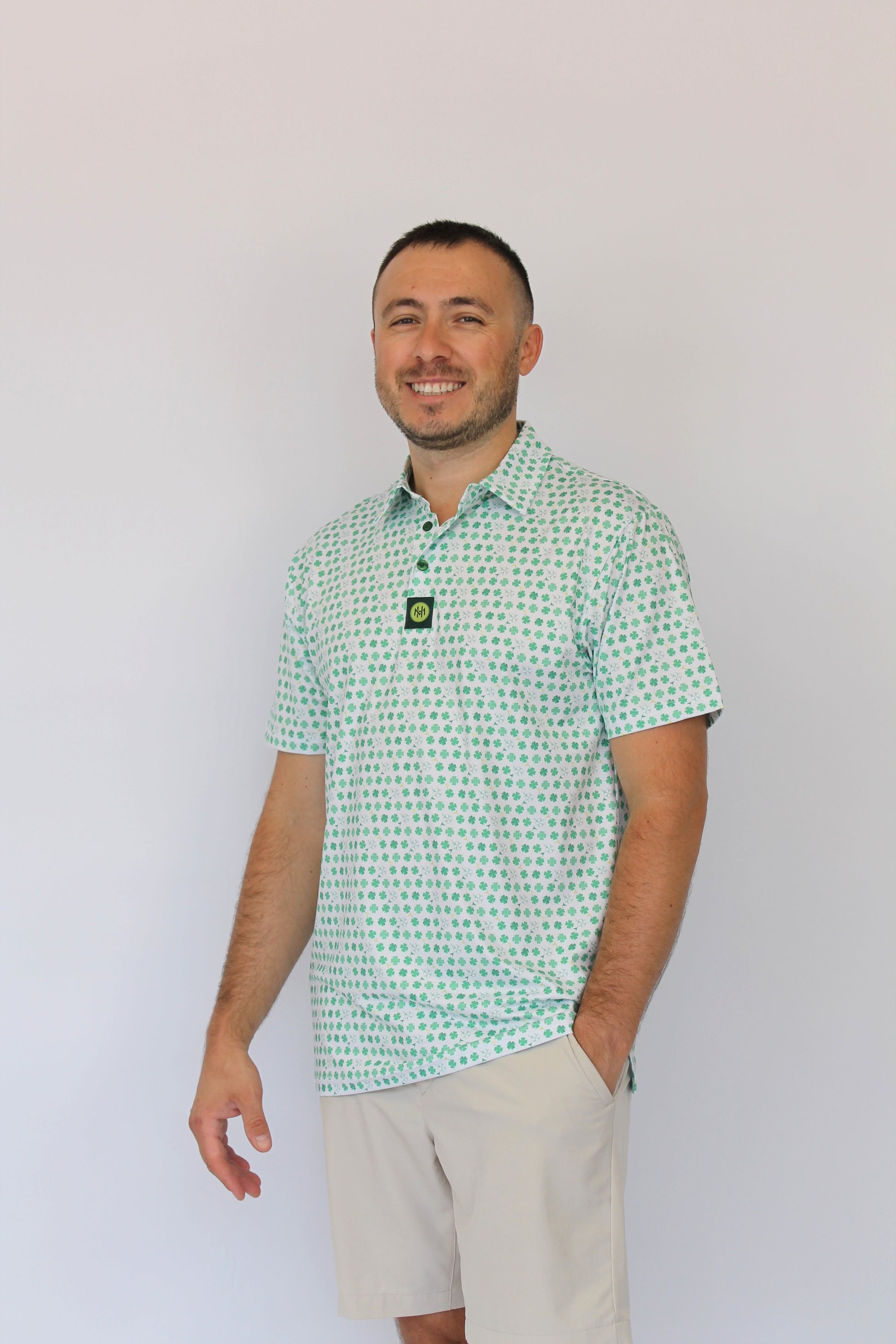 A man wearing a light green and white Clover patterned short-sleeve polo shirt with beige shorts, standing against a plain white background.