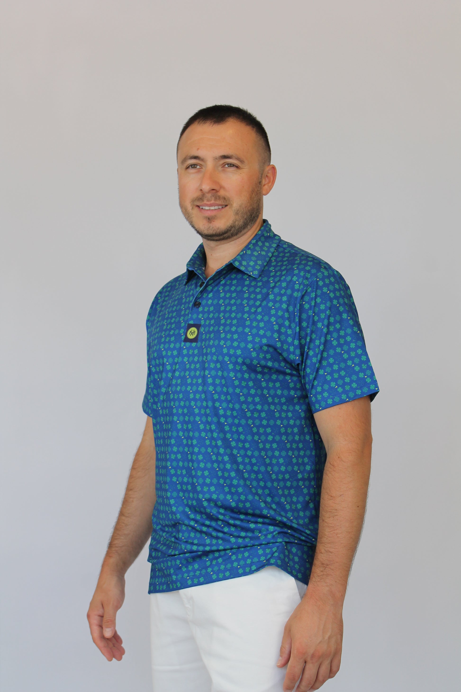 A man wearing the Amen Clover Golf Polo blue short-sleeve polo shirt with a small yellow clover pattern and white pants, shown against a plain light gray background.