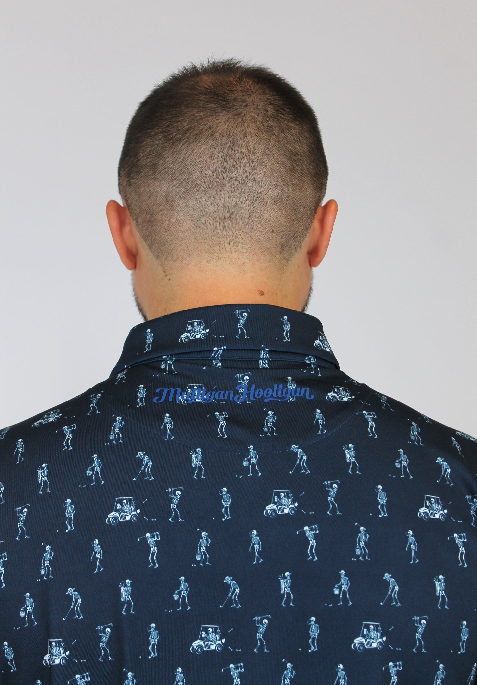 Back view of a man wearing a dark navy blue collared shirt with a light blue golfing skeletons golf-themed all-over print, featuring golfers and carts. The brand name 'Mulligan Hooligan' is visible in script on the inner neck label.
