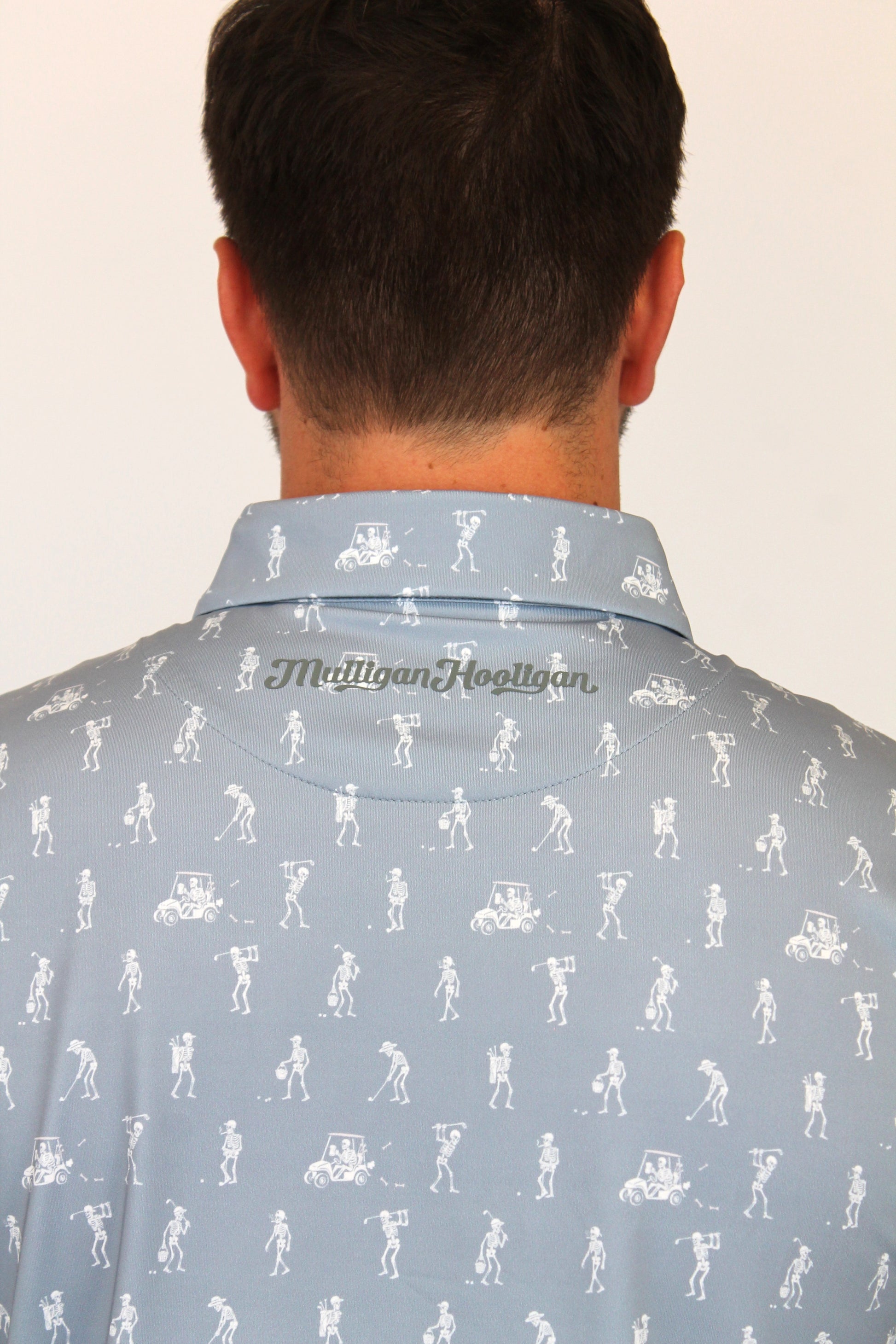 Back view of a man wearing a light grey collared shirt with white skeletons playing golf all-over print and the 'Mulligan Hooligan.' brand name printed on the inner neck label.