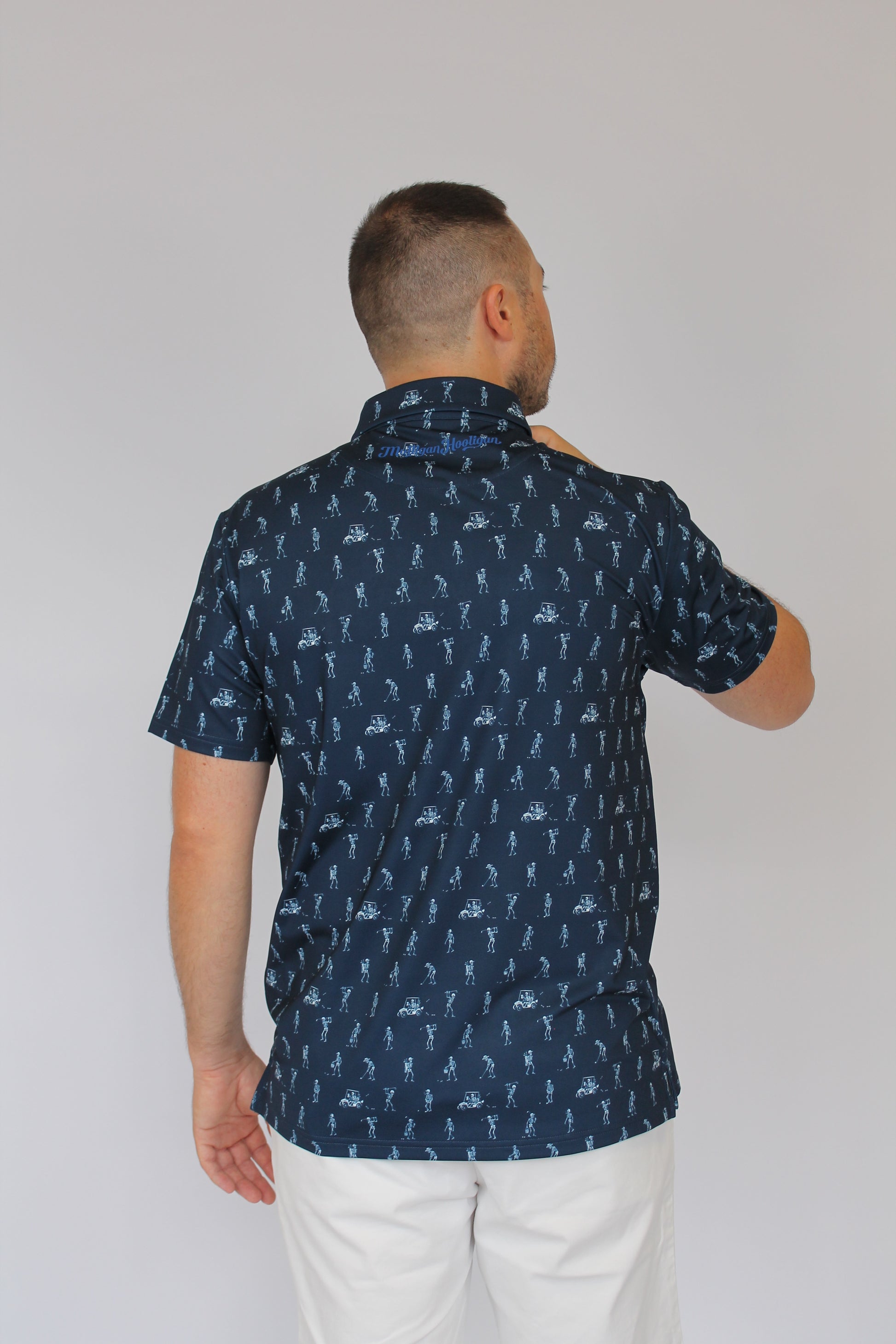 A man wearing a navy blue short-sleeve button-up shirt with a repeating white golfing skeletons pattern, paired with white pants, shown from a side profile against a plain light gray background.