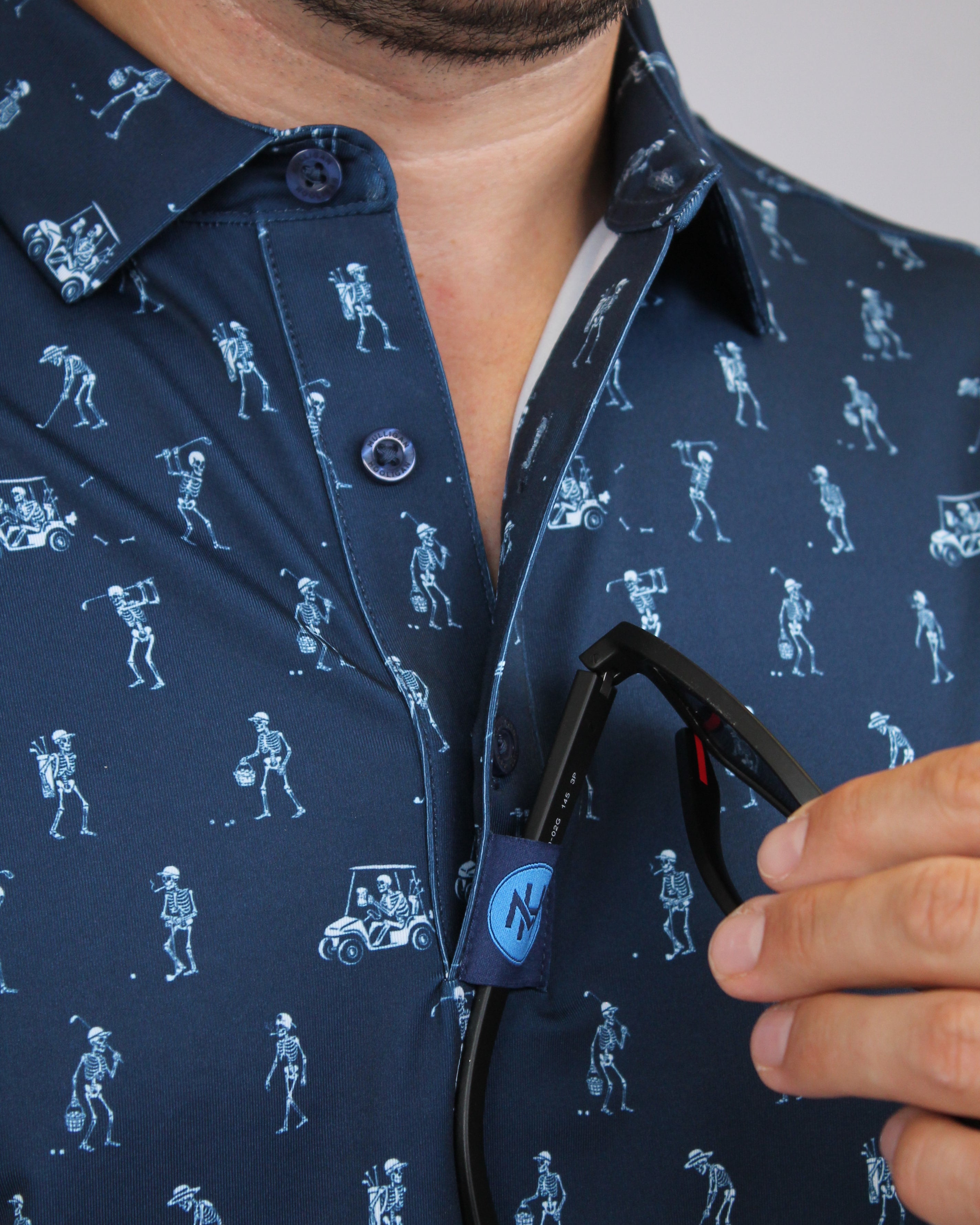 Close-up of a man wearing a navy blue golfing skeletons patterned shirt and holding his sunglasses on a placket custom loop. 