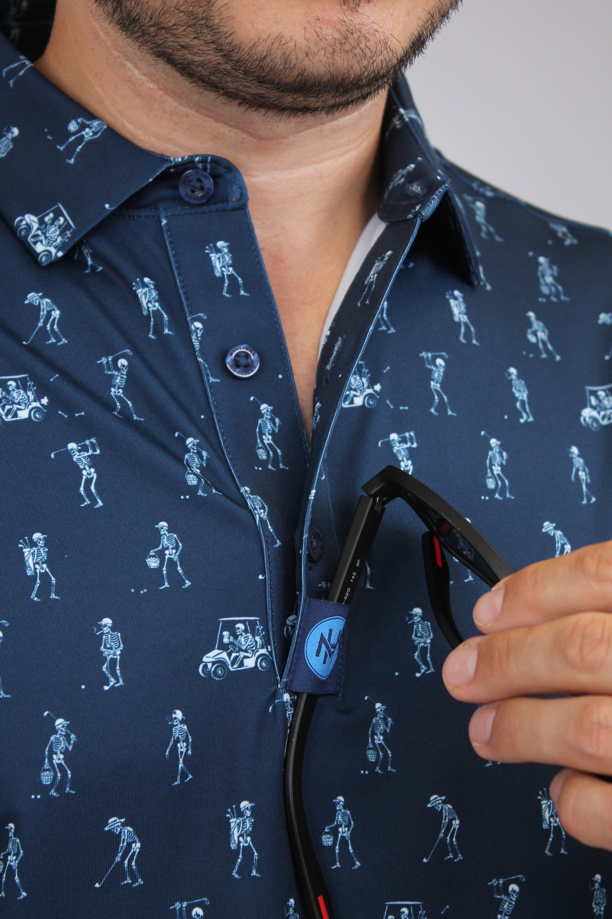 Close-up of a man wearing a navy blue golfing skeletons patterned shirt and holding his sunglasses on a placket custom loop. 
