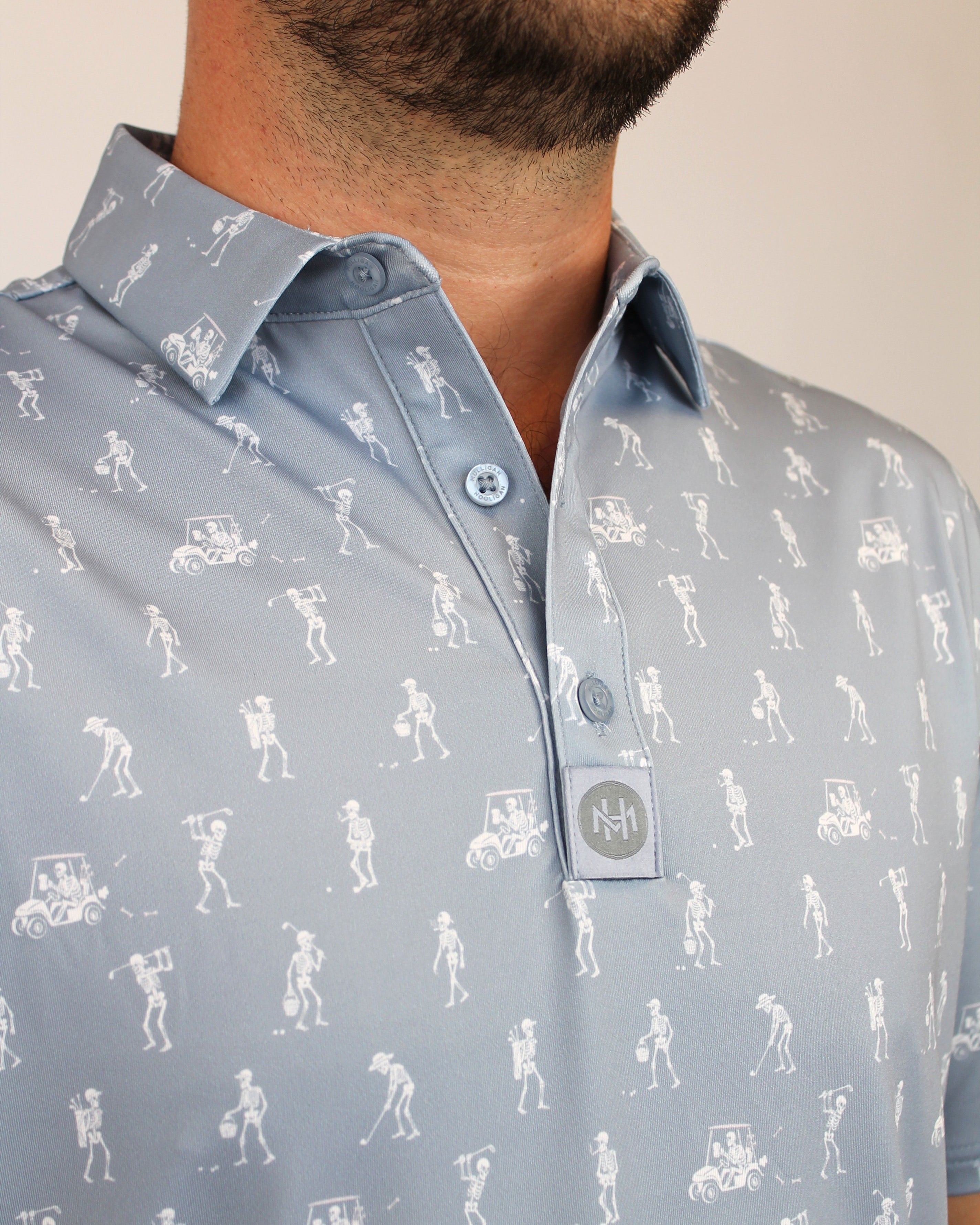 A close-up of a man wearing a light gray, silver polo shirt with a white all-over golf-themed skeletons playing golf graphic print, featuring golf clubs and players.