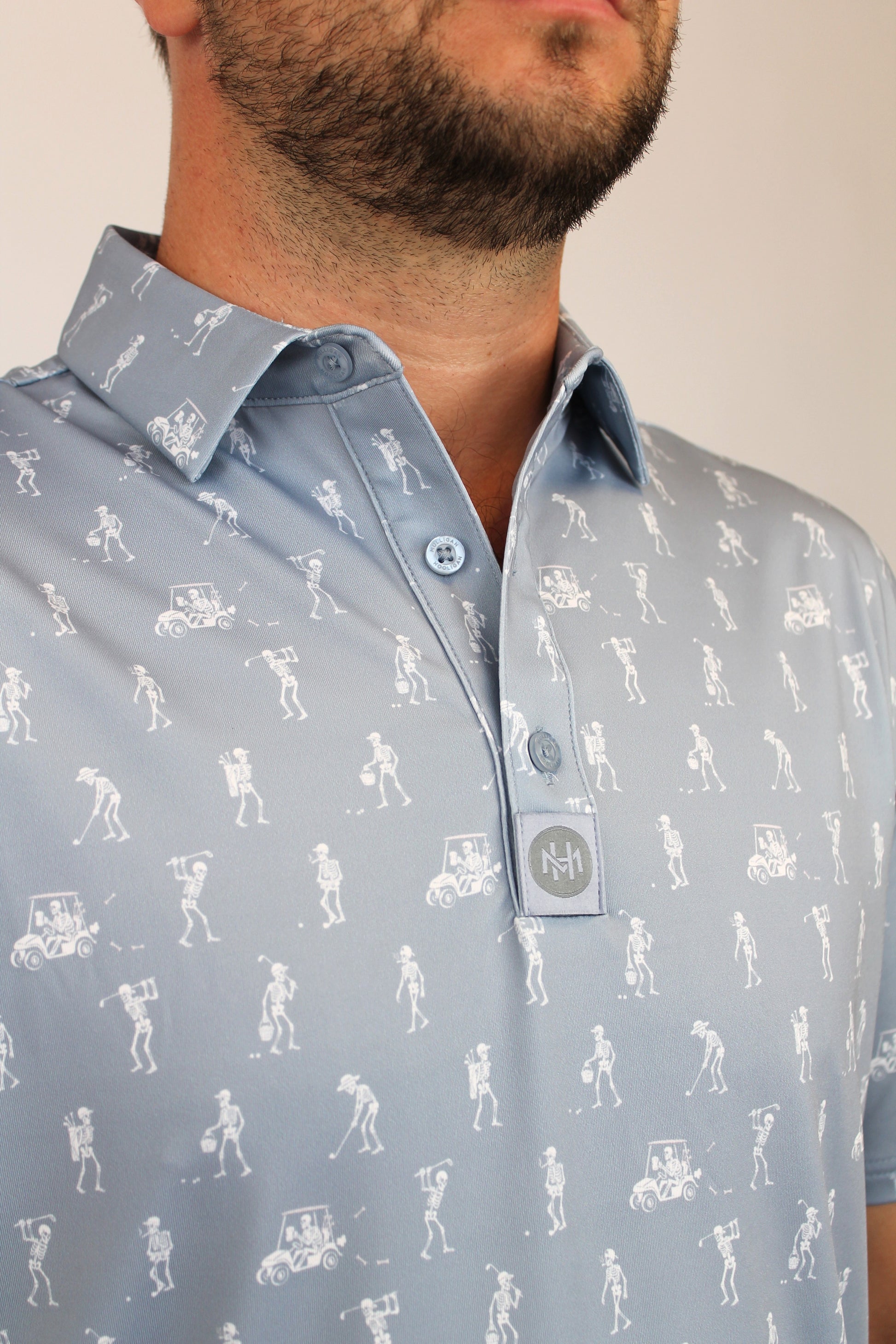 A close-up of a man wearing a light gray, silver polo shirt with a white all-over golf-themed skeletons playing golf graphic print, featuring golf clubs and players.