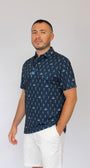 A man wearing a navy blue short-sleeve button-up shirt with a white skeletons playing golf on a golf course pattern, paired with white shorts, standing against a light gray studio background.