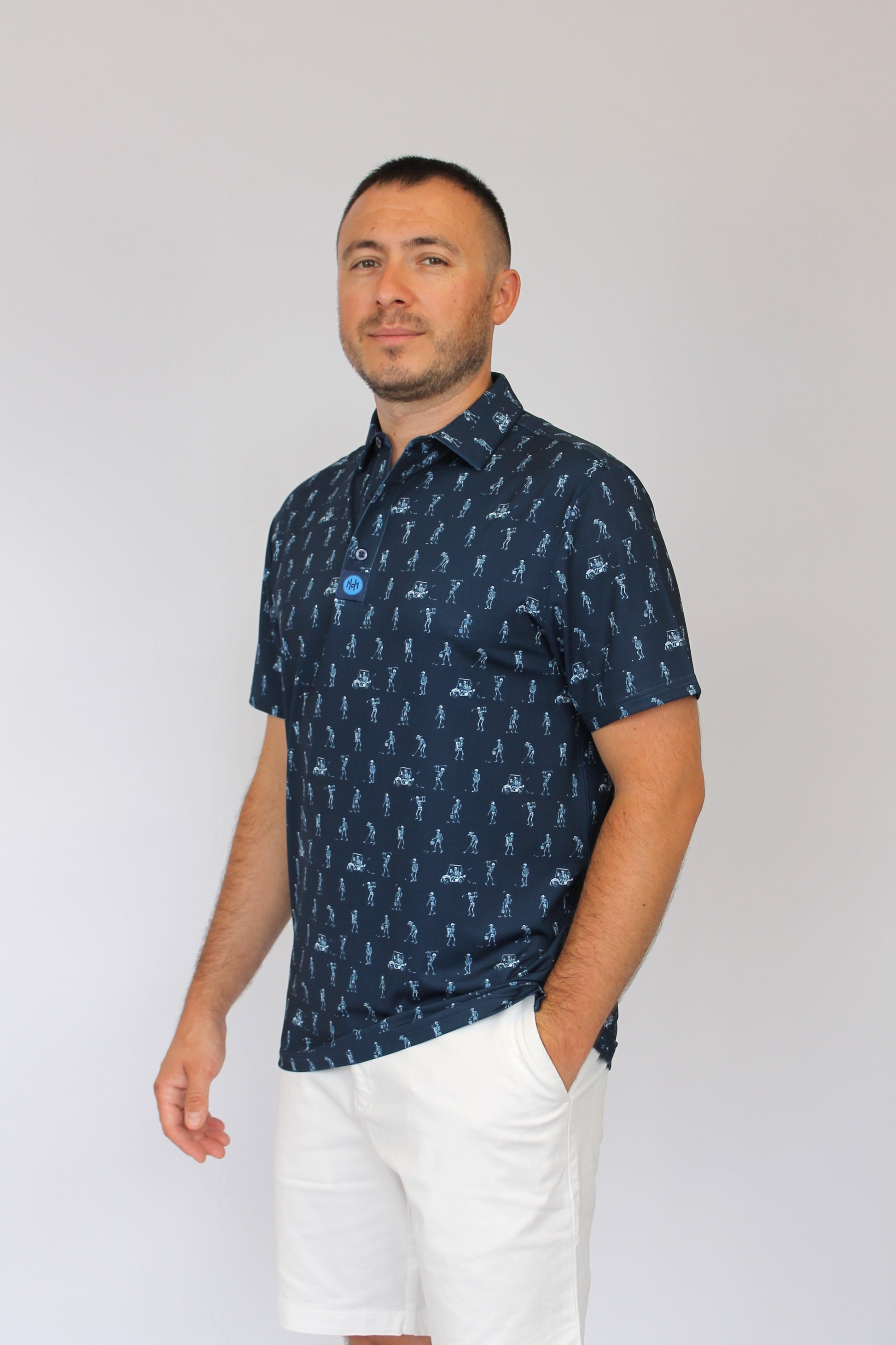 A man wearing a navy blue short-sleeve button-up shirt with a white skeletons playing golf on a golf course pattern, paired with white shorts, standing against a light gray studio background.