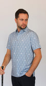 A man wearing a light blue short-sleeve polo shirt with white golfing skeletons on a golf course pattern, holding a golf club against a plain light gray background.
