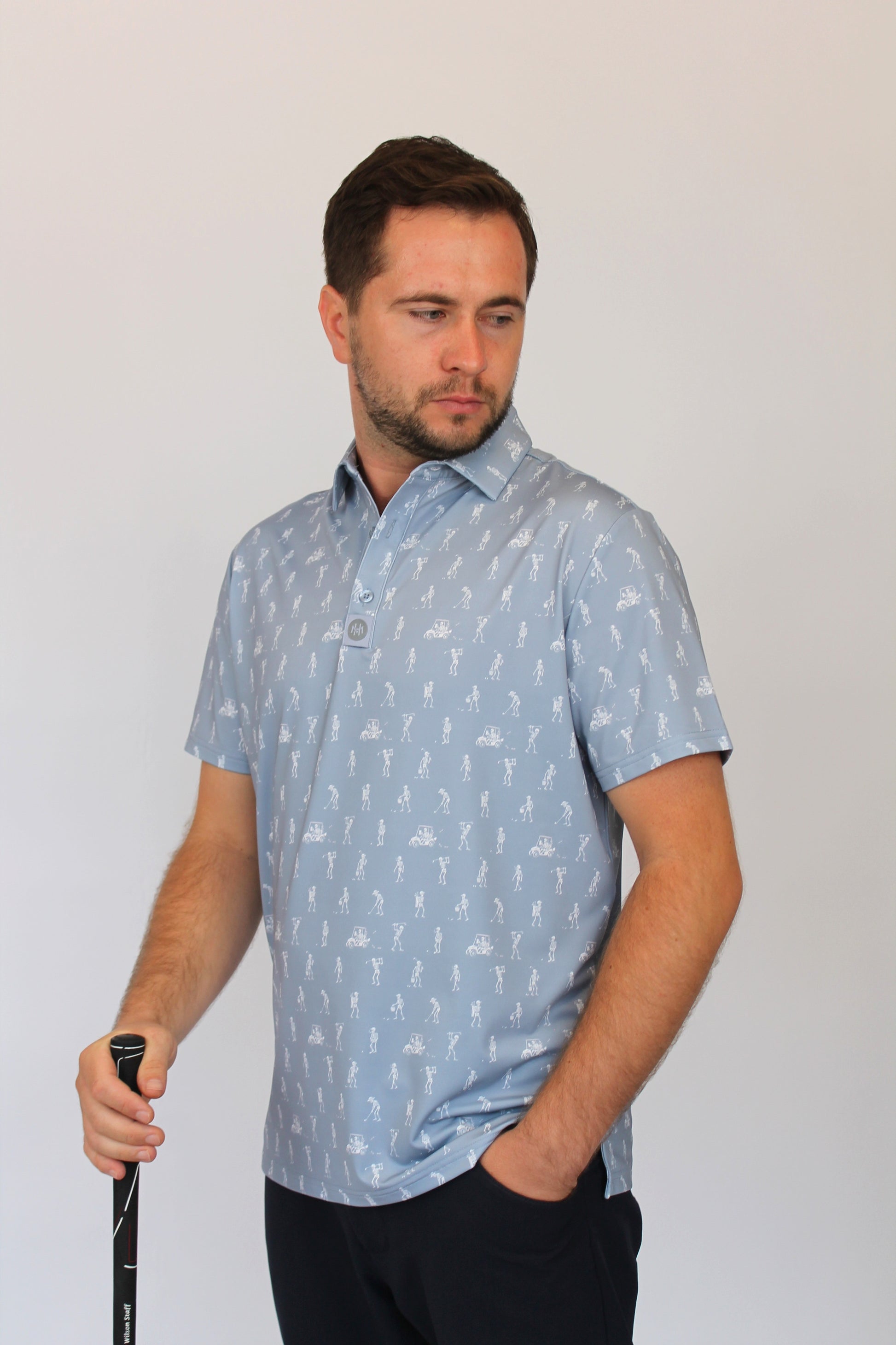 A man wearing a light blue short-sleeve polo shirt with white golfing skeletons on a golf course pattern, holding a golf club against a plain light gray background.