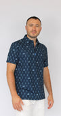 A man wearing a navy blue short-sleeve button-up shirt with a white golfing skeletons pattern, paired with white pants, standing against a plain light gray background.