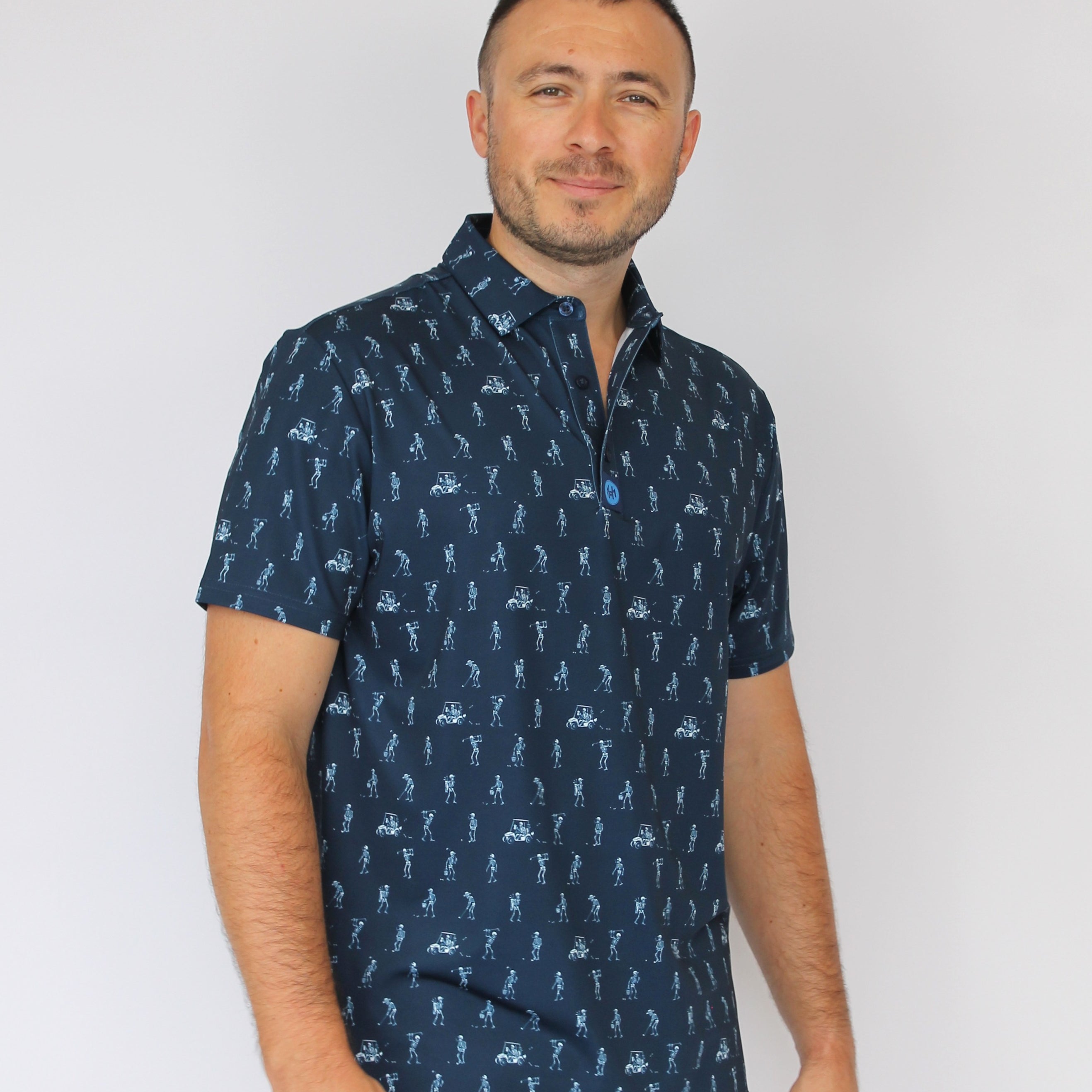 A man wearing a navy blue short-sleeve button-up shirt with a white golfing skeletons pattern, paired with white pants, standing against a plain light gray background.