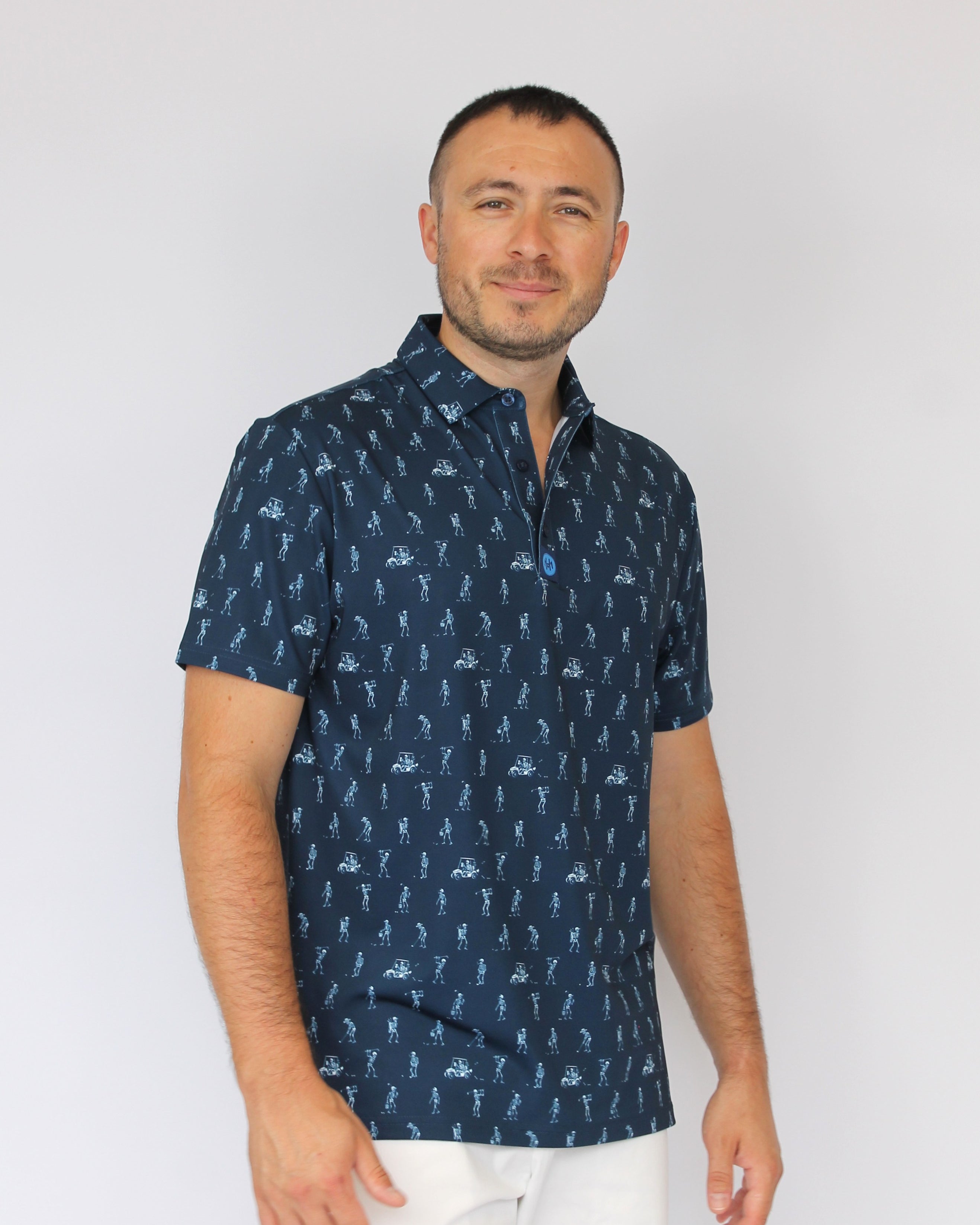 A man wearing a navy blue short-sleeve button-up shirt with a white golfing skeletons pattern, paired with white pants, standing against a plain light gray background.