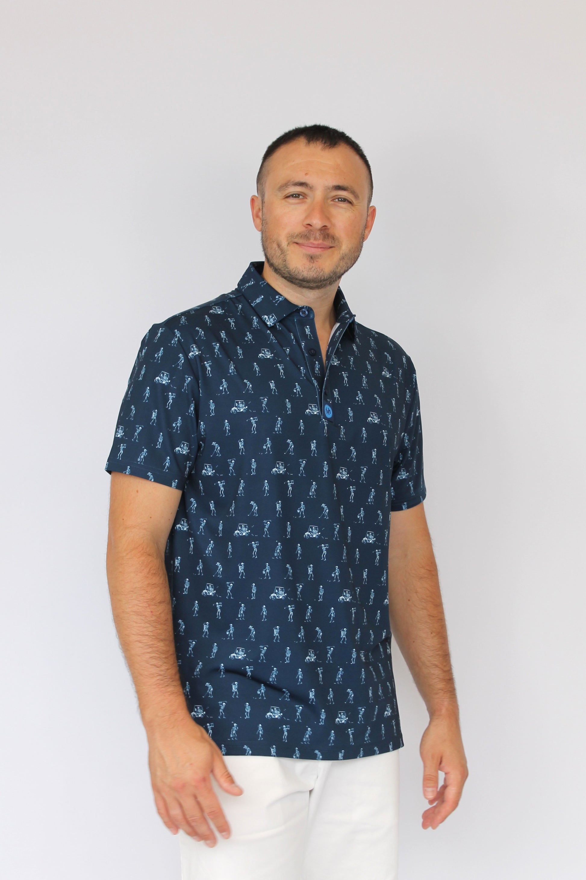 A man wearing a navy blue short-sleeve button-up shirt with a white golfing skeletons pattern, paired with white pants, standing against a plain light gray background.