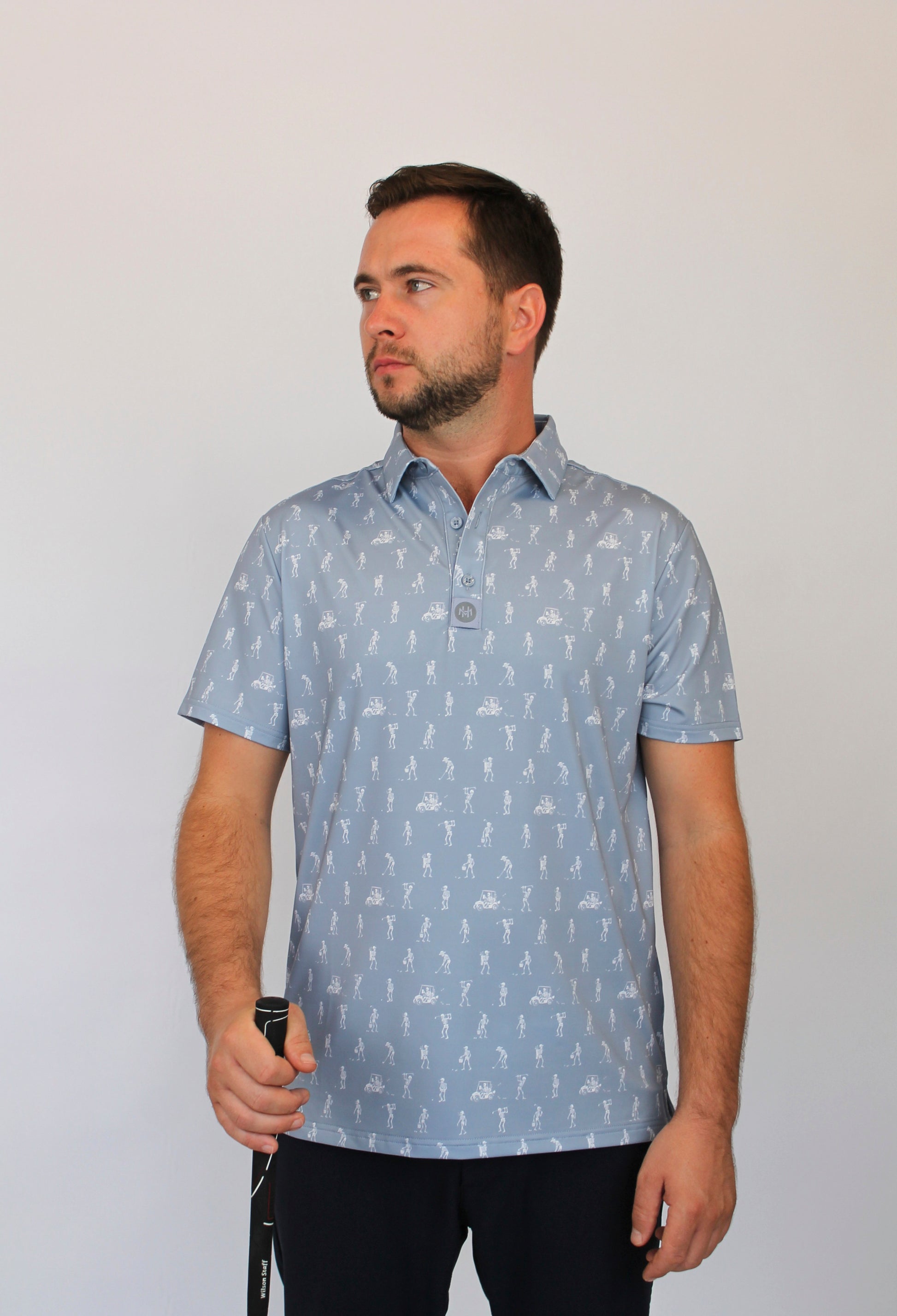 A man wearing a light gray short-sleeve polo shirt with white goofing skeletons pattern, standing against a plain light gray background.