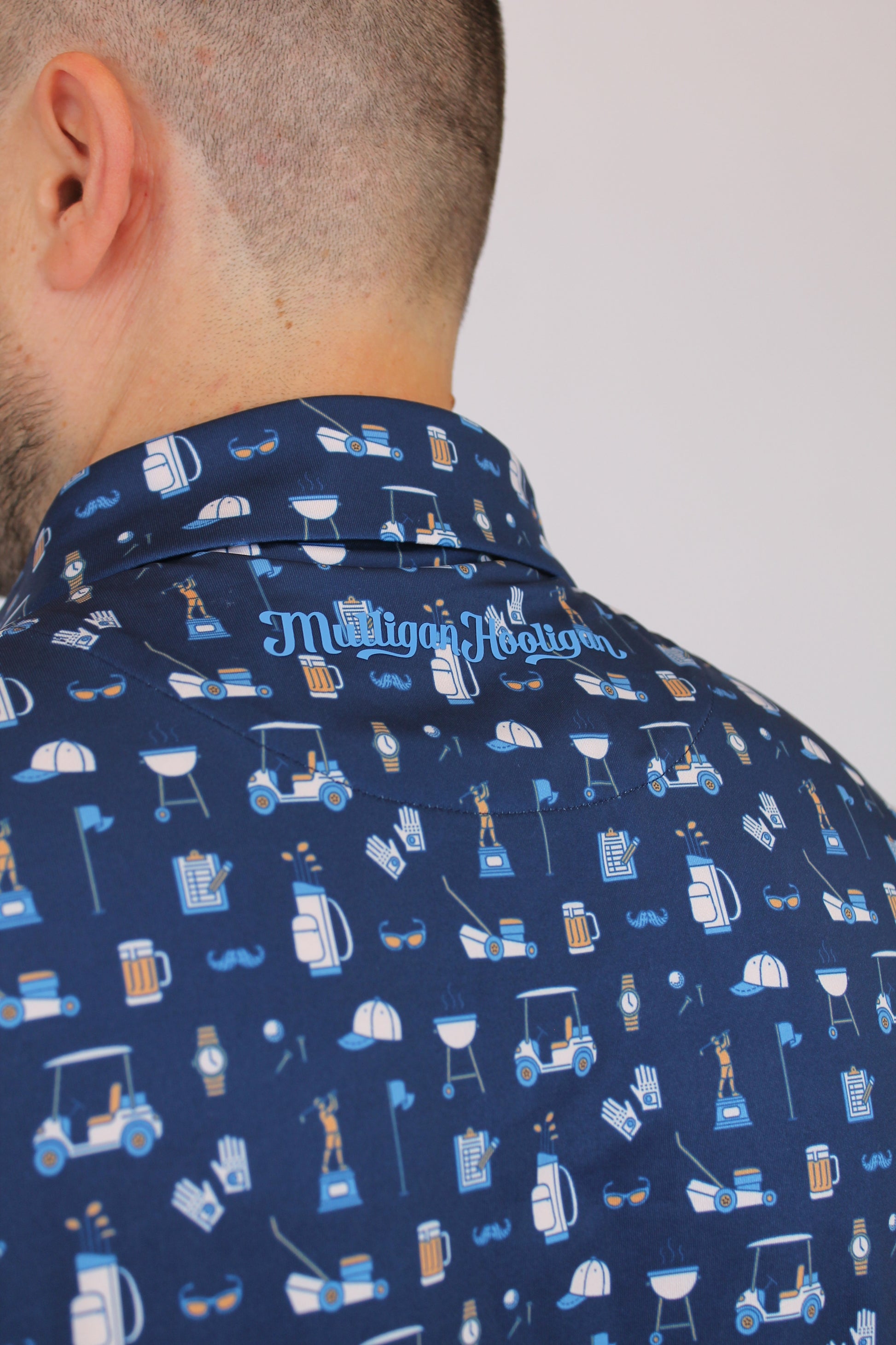 Close-up of a person wearing a navy blue Golf Dad Polo with a repeating pattern of golf clubs and a script of 'Mulligan Hooligan' logo.
