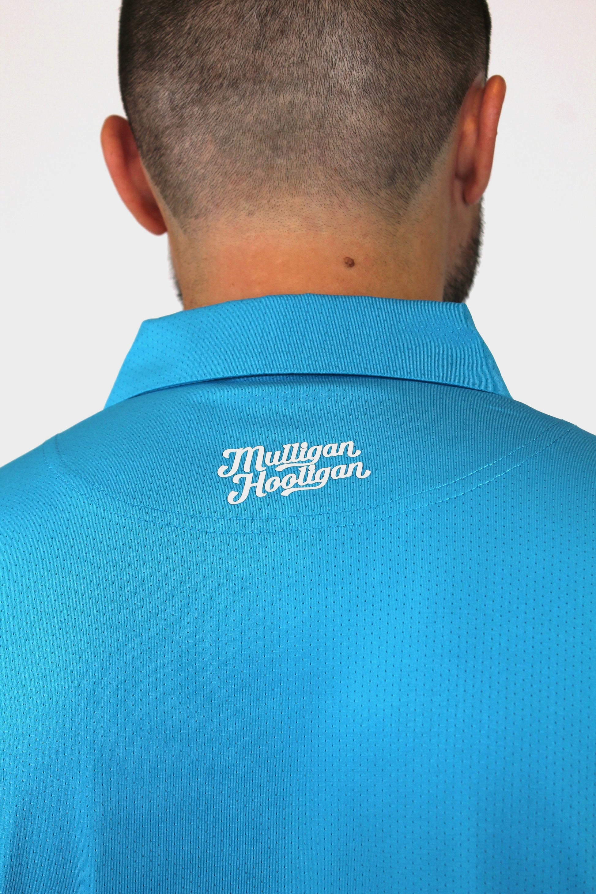 Back view of a man wearing a bright blue collared polo shirt with 'Mulligan Hooligan' embroidered in white on the upper back, against a plain light background.
