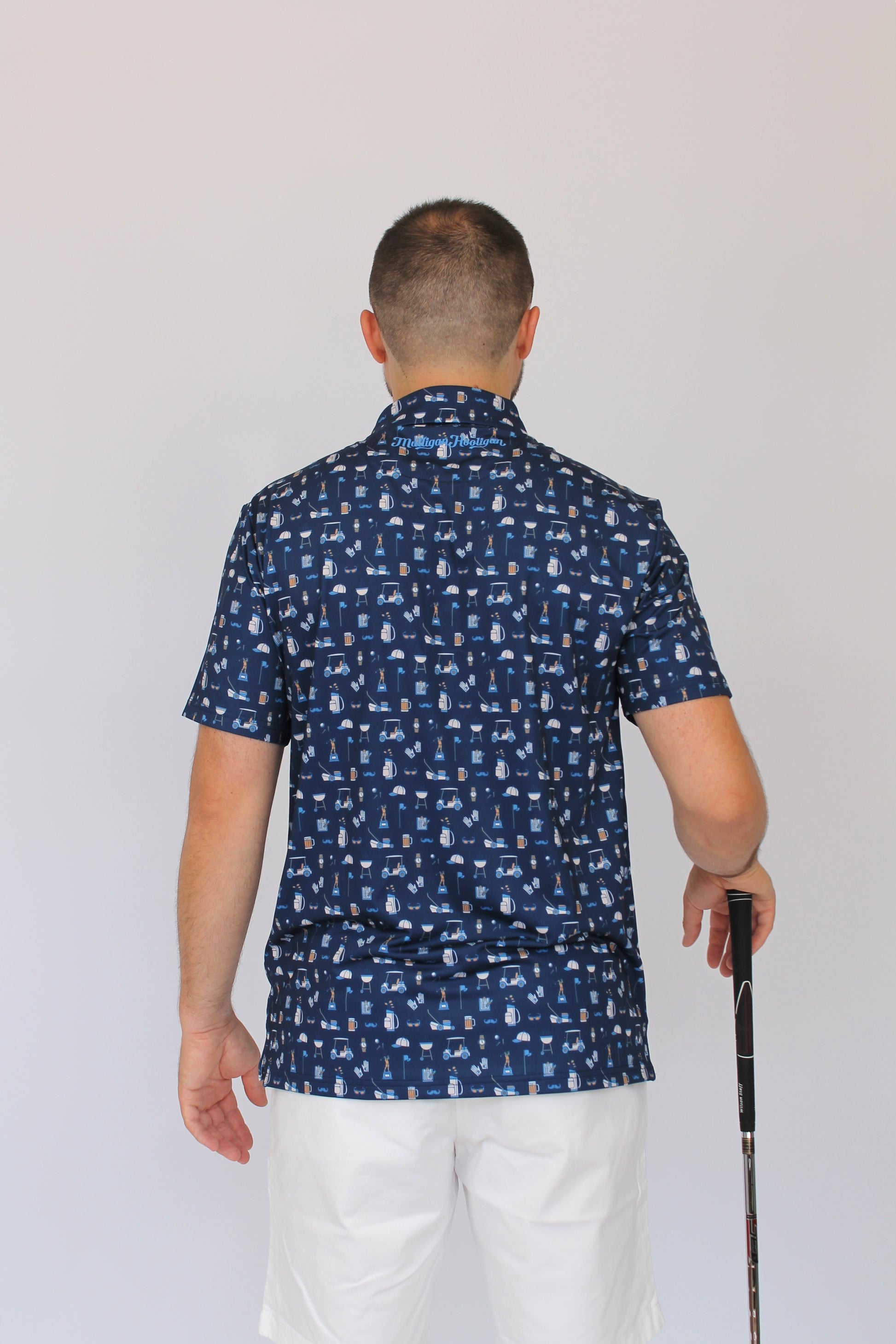 A man viewed from behind wearing a navy blue short-sleeve button-up shirt with a white golf dad-themed pattern, paired with white pants and holding a golf club against a plain light gray background.