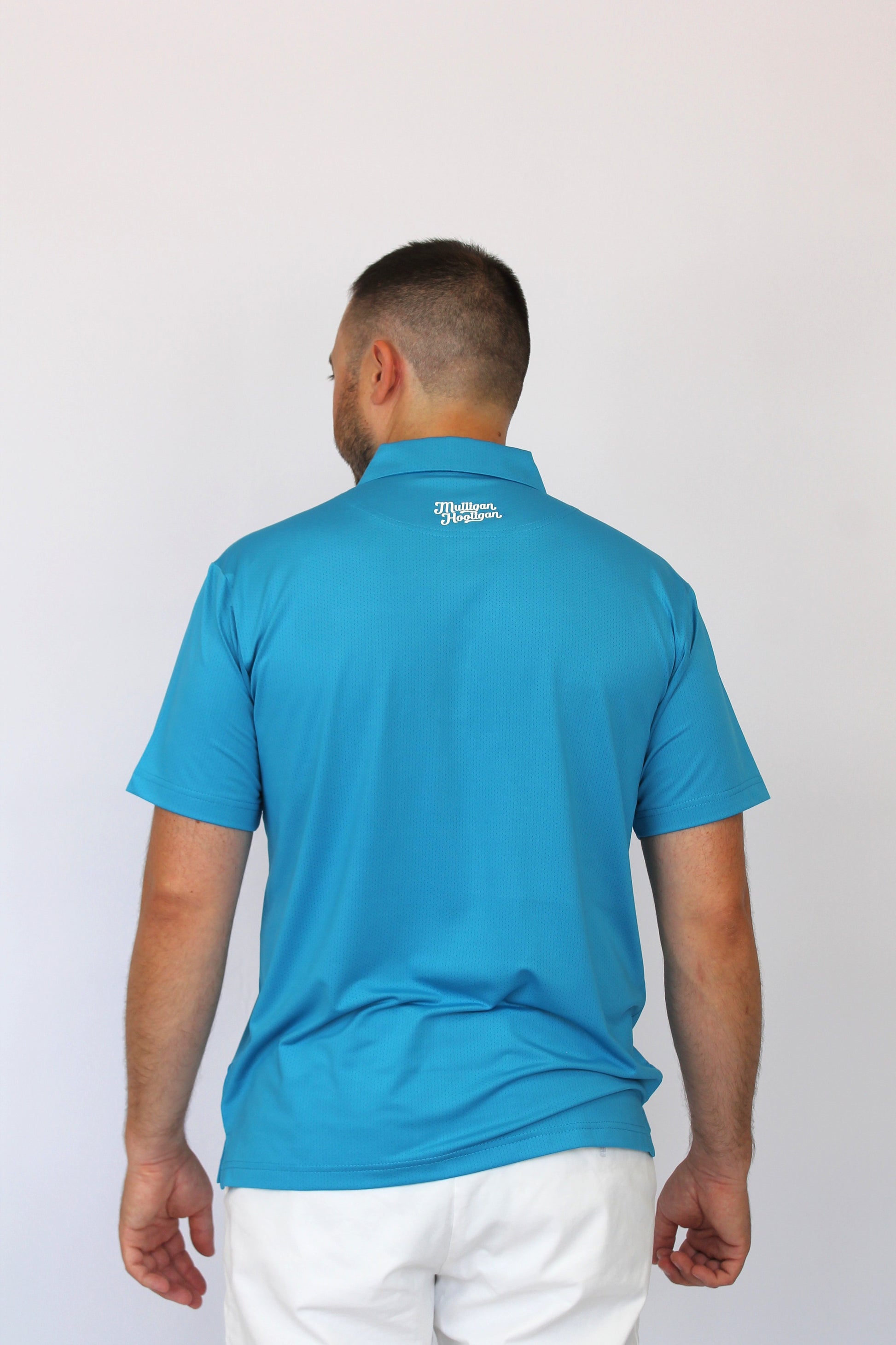 A man viewed from behind wearing a bright turquoise blue short-sleeve polo shirt and white pants, standing against a plain light gray background.