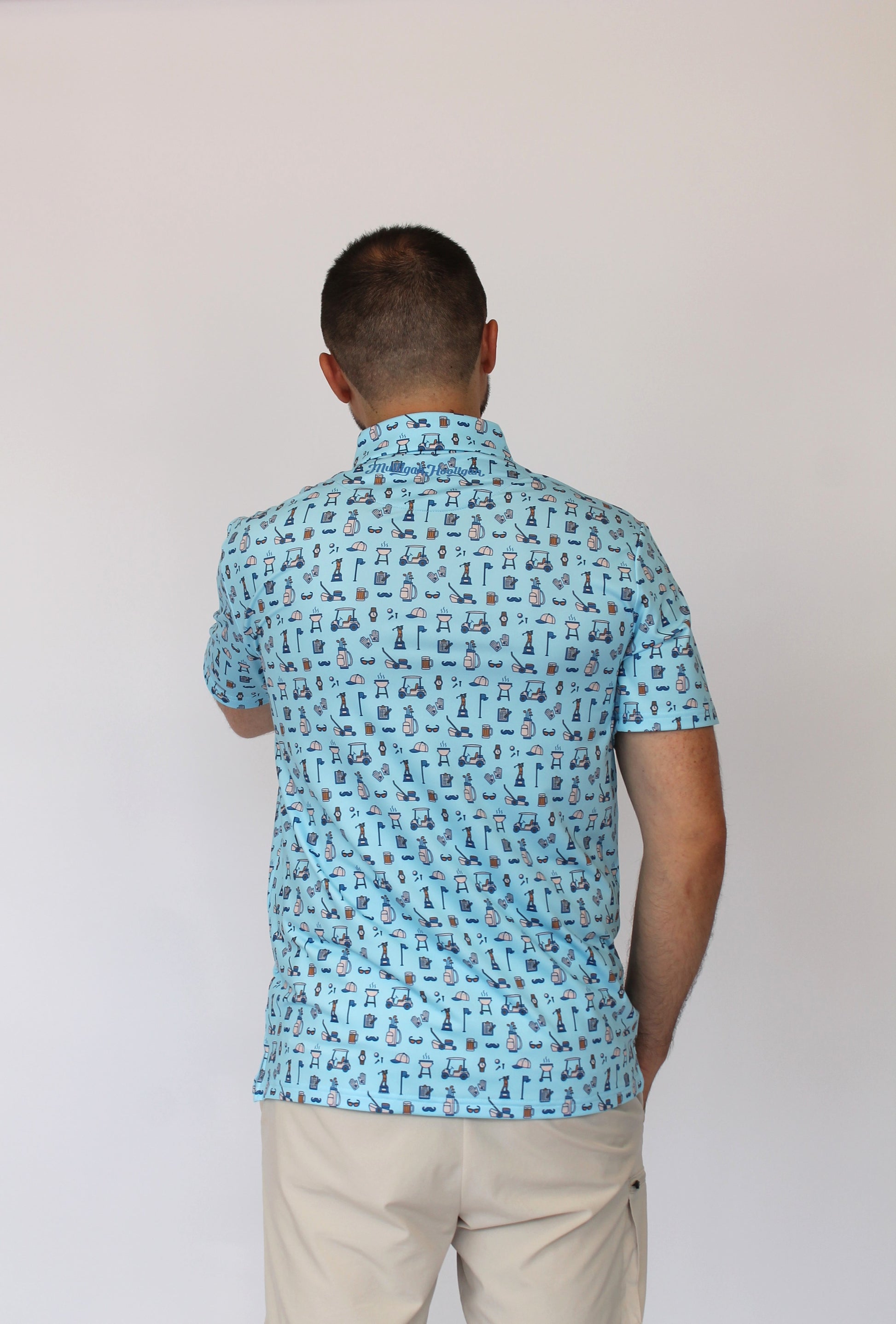A man viewed from behind wearing a light blue short-sleeve button-up shirt with a dense black Golfing dad-themed print, paired with beige trousers, against a plain off-white background.