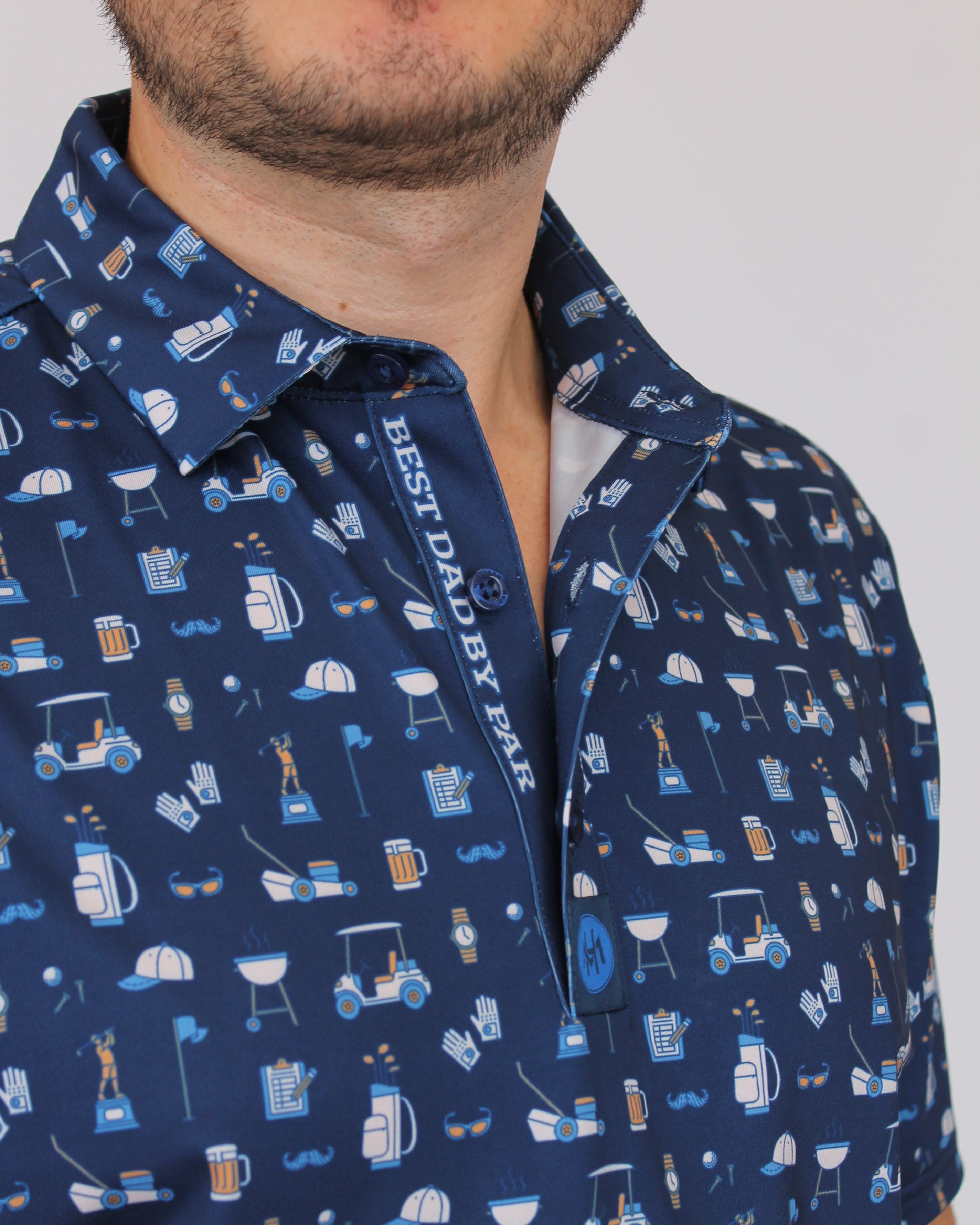 A close-up of a man wearing a navy blue short-sleeve button-up shirt with a white and light blue pattern of household items and the text 'BEST DAD BY PAR' on the inner placket.