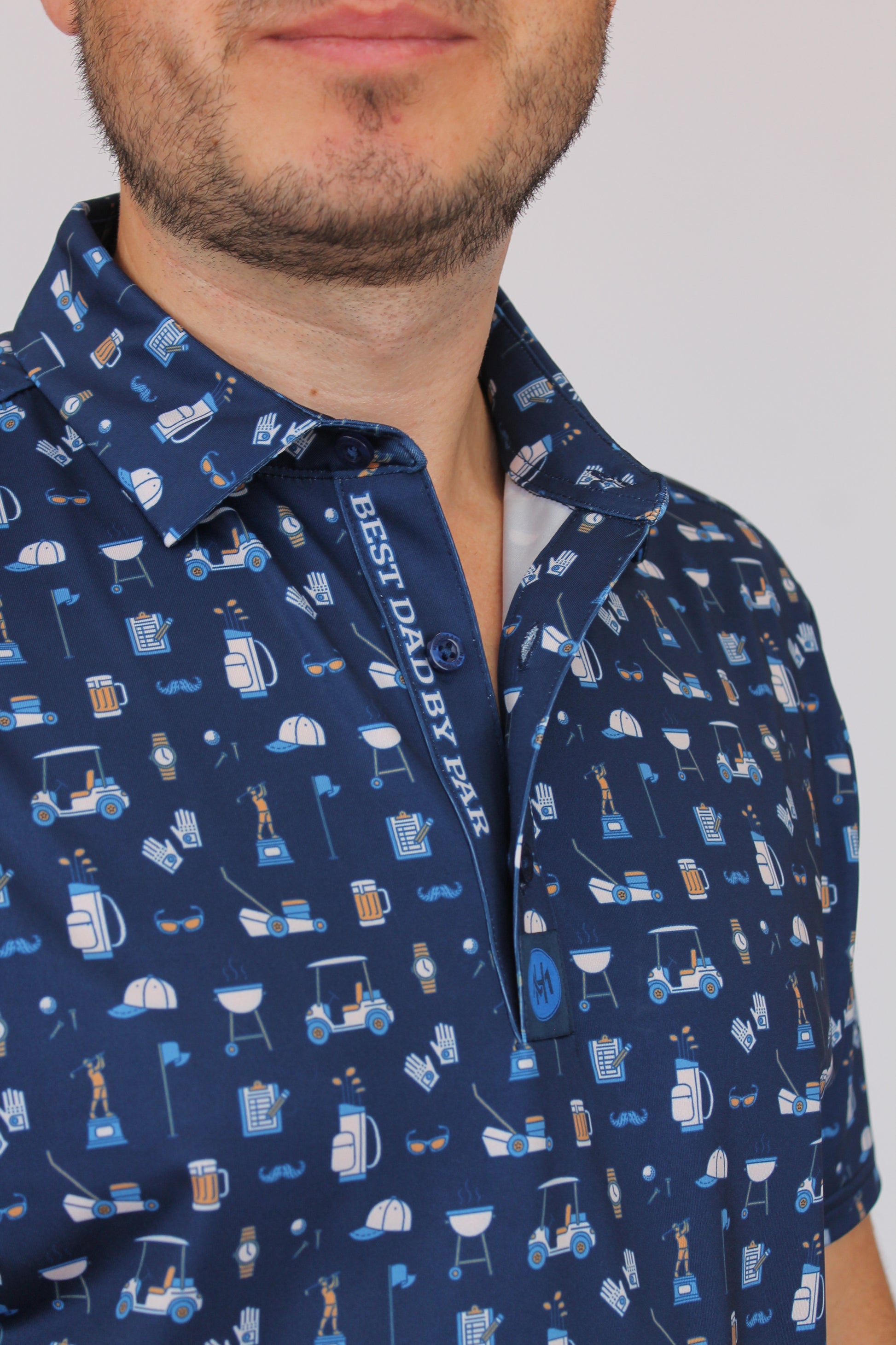 A close-up of a man wearing a navy blue short-sleeve button-up shirt with a white and light blue pattern of household items and the text 'BEST DAD BY PAR' on the inner placket.