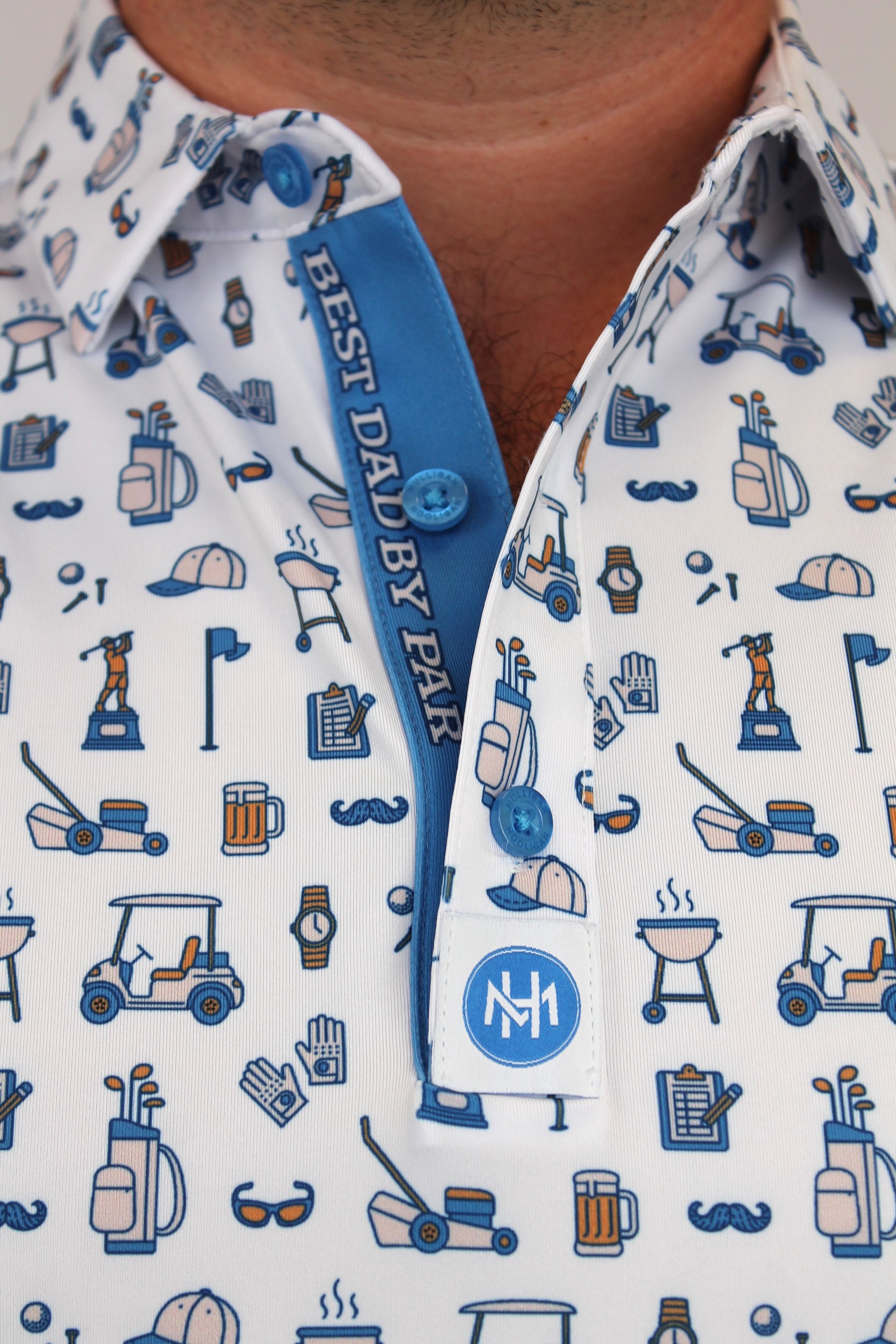 Close-up of a man wearing a white button-down shirt with a blue and navy Dad golf-themed print, featuring a collar, and a custom planchet saying ''Best Dad By Par'' blue placket, and a white chest patch with a circular 'NM' logo.