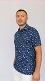 A man wearing a navy blue Best Dad By Par short-sleeve button-up shirt with a white Dad pattern of golf carts, grills, mustaches, dad, and beers, paired with white pants, standing against a plain light gray background.