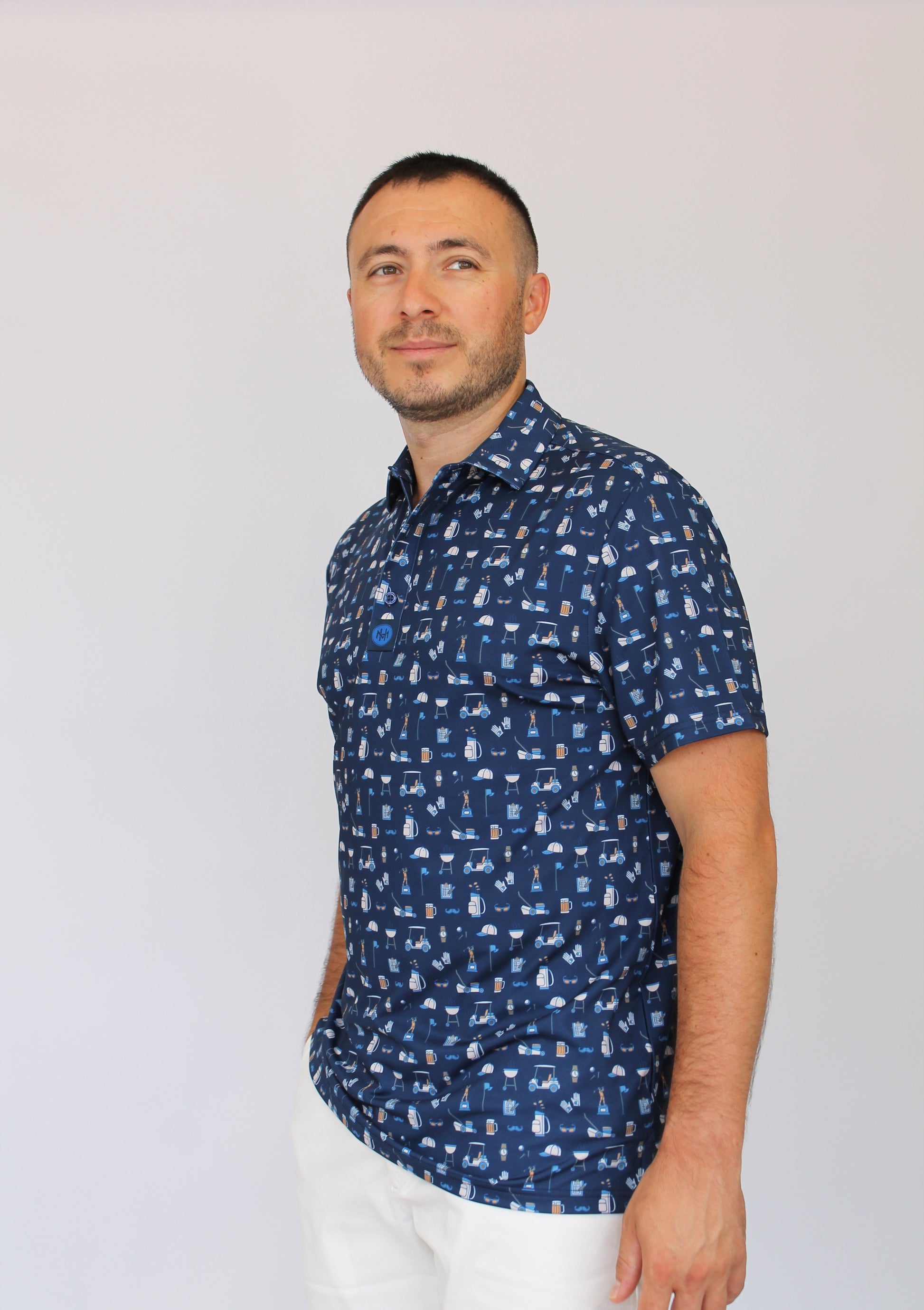 A man wearing a navy blue Best Dad By Par short-sleeve button-up shirt with a white Dad pattern of golf carts, grills, mustaches, dad, and beers, paired with white pants, standing against a plain light gray background.