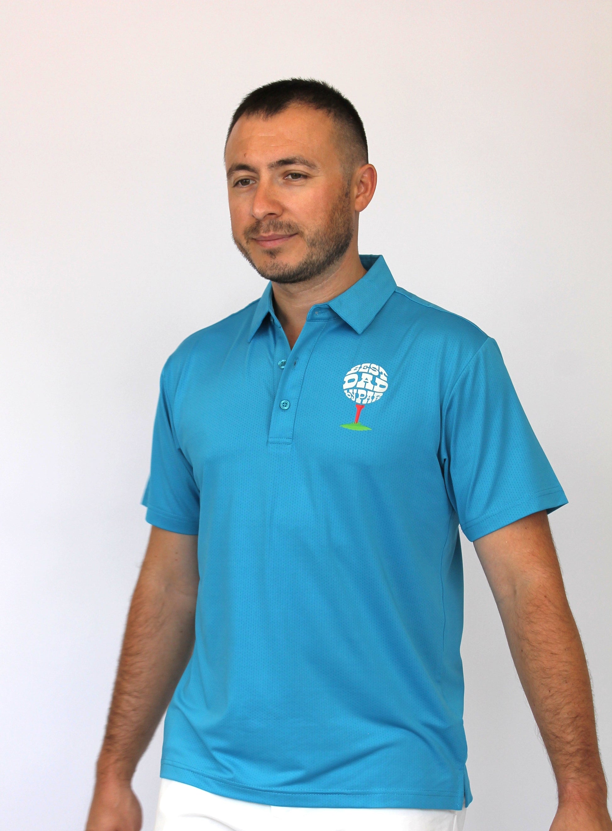 A man wearing a bright blue polo shirt with a golf-themed logo on the left chest that says ''BEST DAD BY PAR'', paired with white pants, standing against a plain light background.