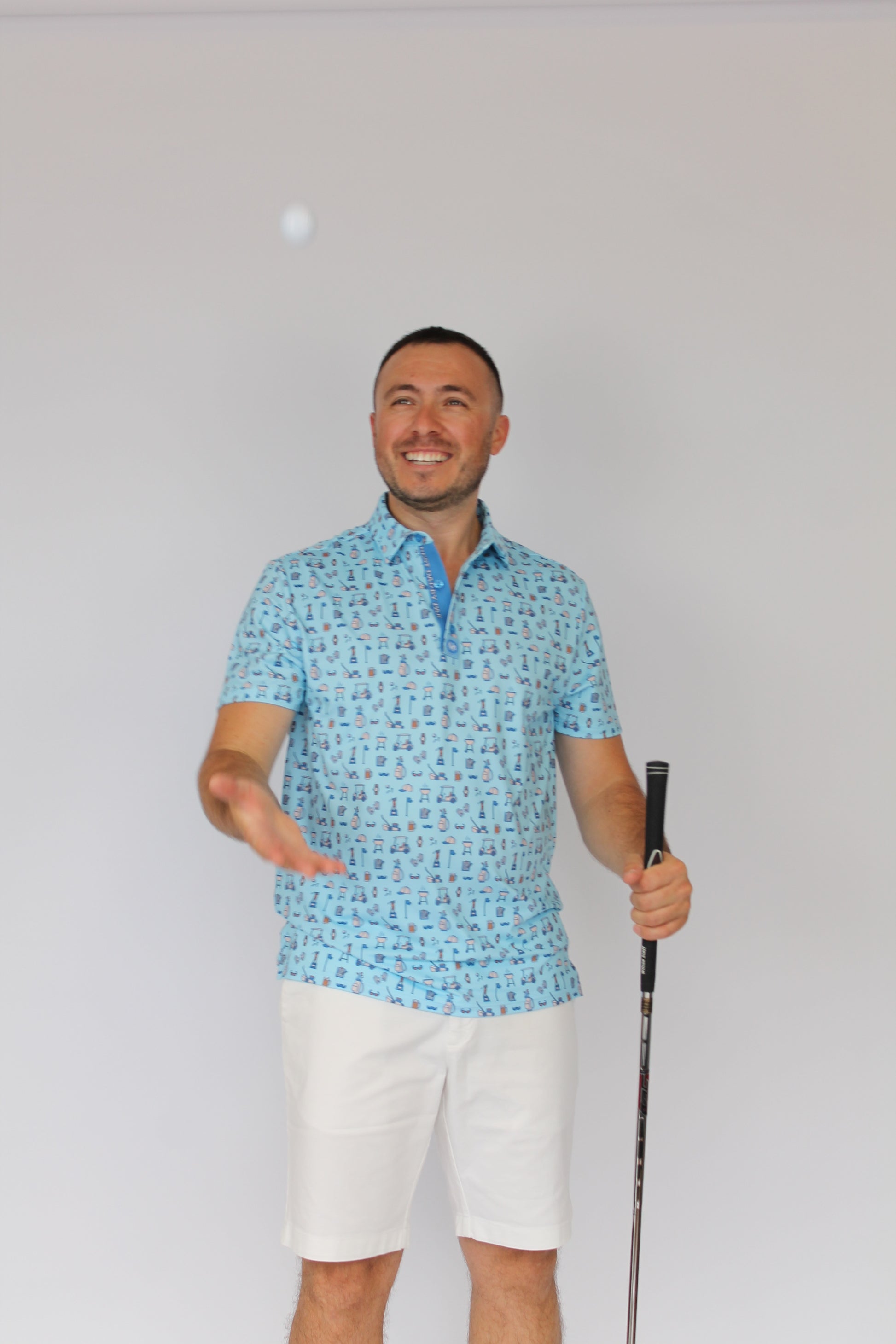 A man wearing a light blue patterned Best Dad By Par short-sleeve polo shirt and white shorts, holding a golf club and smiling against a plain white background.
