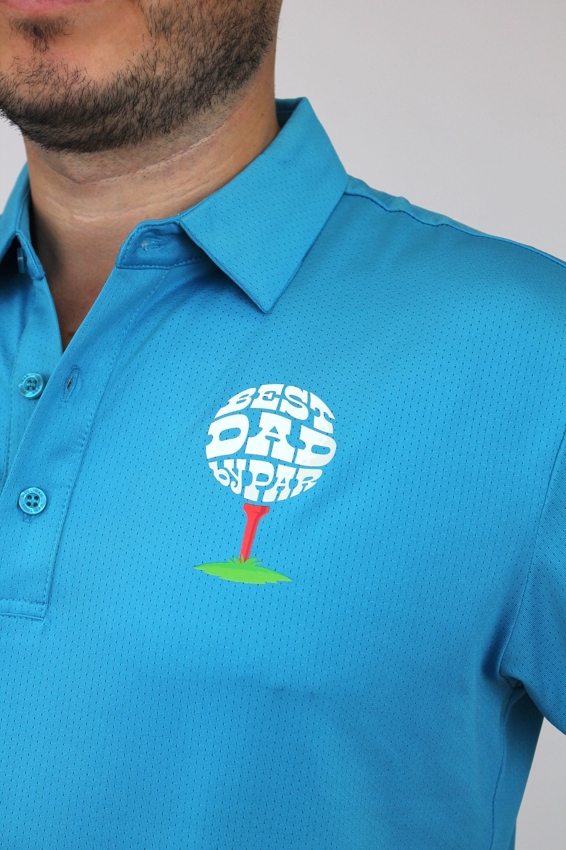A close-up of a man wearing a bright blue polo shirt with a custom golf-themed chest graphic. The graphic features a white golf ball with the words 'BEST DAD BY PAR' in white, resting on a red tee over a green patch of grass.