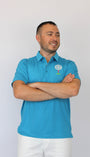 A man wearing a bright blue polo shirt with a small white and green logo on the left chest, paired with white pants, standing against a plain light gray background with his arms crossed.